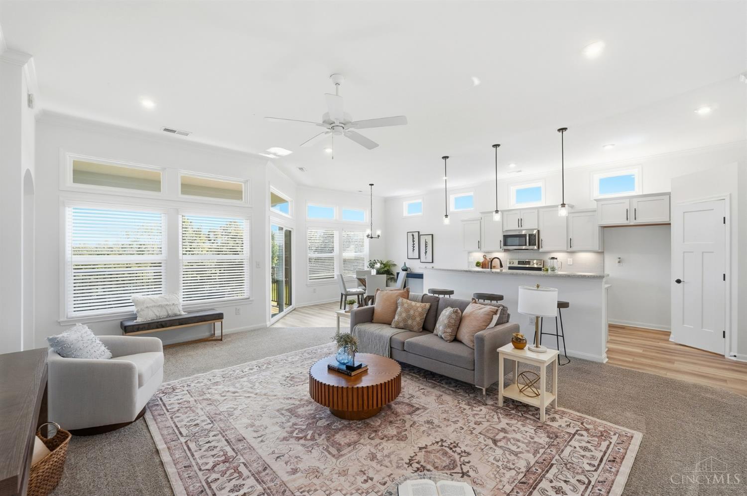 Furnished living room opens to a white kitchen, with large windows, high ceilings, and a patterned rug.