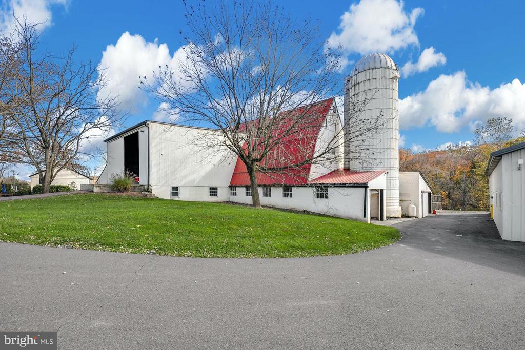 White farm building with red roof and tall silo beside a paved driveway and lawn.