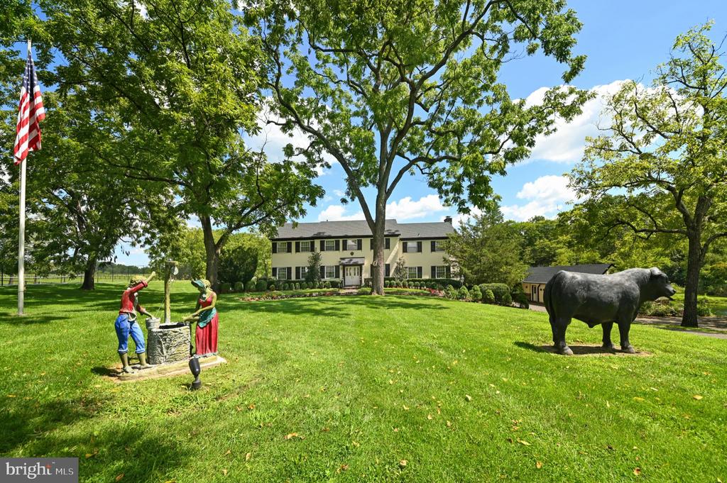 Two-story house behind a broad lawn with mature trees, a stone well, figurines, and a horse sculpture.