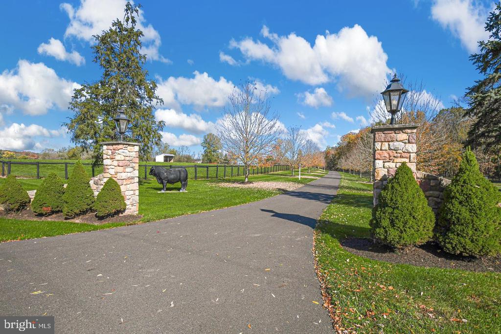 Paved rural property entrance with stone lantern pillars, clipped shrubs, black fencing, and a cow statue.