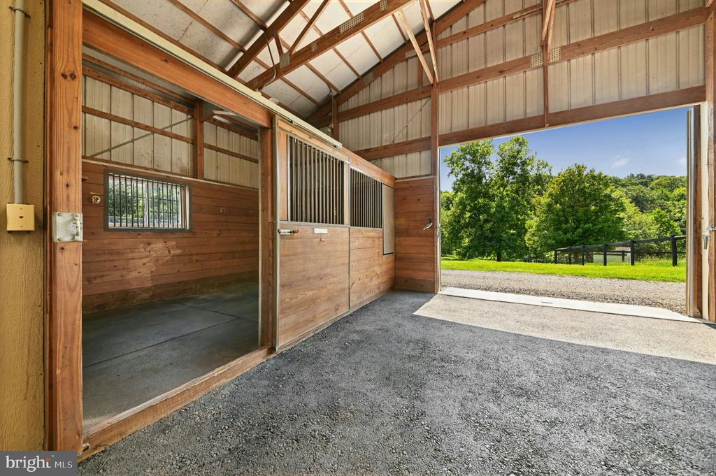 Horse barn interior with wooden stalls and an open doorway facing a sunny lawn and paddock.