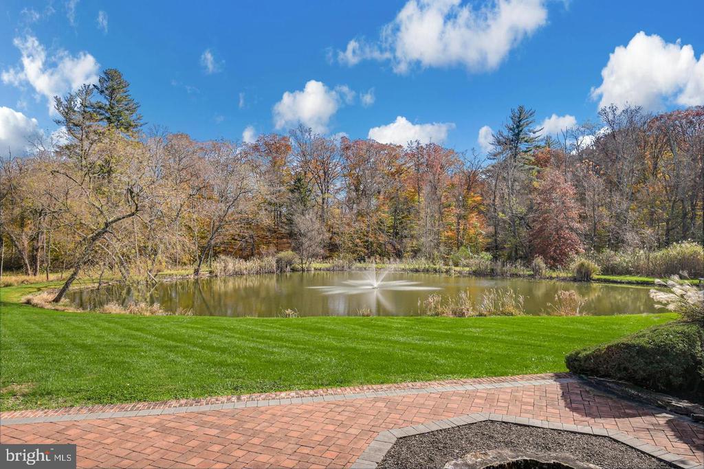 Landscaped pond with a fountain, green lawn, autumn trees, and brick patio.