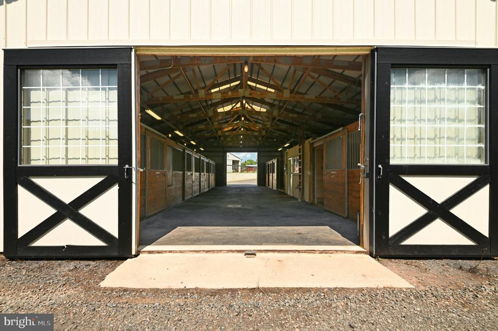 Stable aisle with wooden horse stalls, exposed roof trusses, and open black-and-white barn doors.