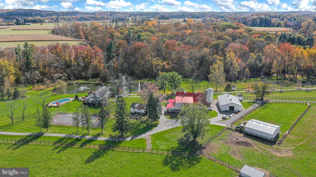 Aerial view of a rural estate with a pool, outbuildings, fenced paddocks, and autumn woodland.