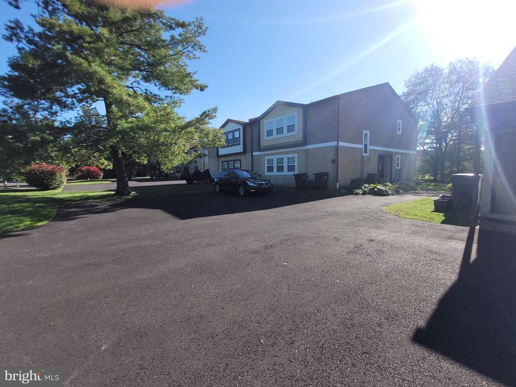 Multi-unit residential building beside a broad paved parking area and mature trees.