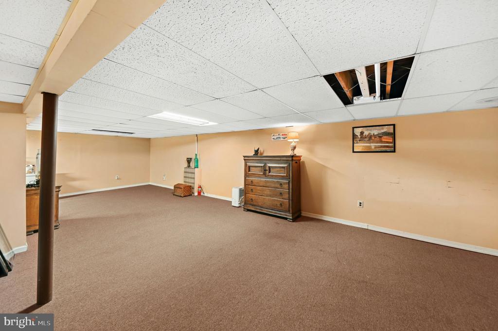 Carpeted basement room with a wooden dresser, support post, and missing ceiling tiles.