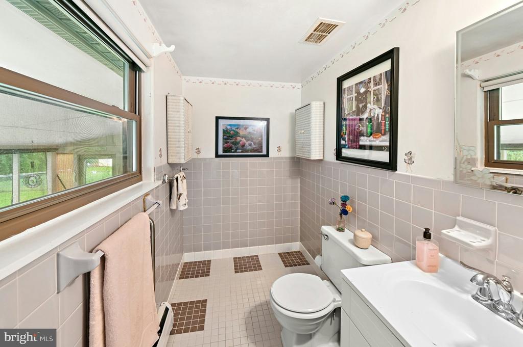 Bathroom with pale pink tile, a white vanity and toilet, large windows, and framed artwork.