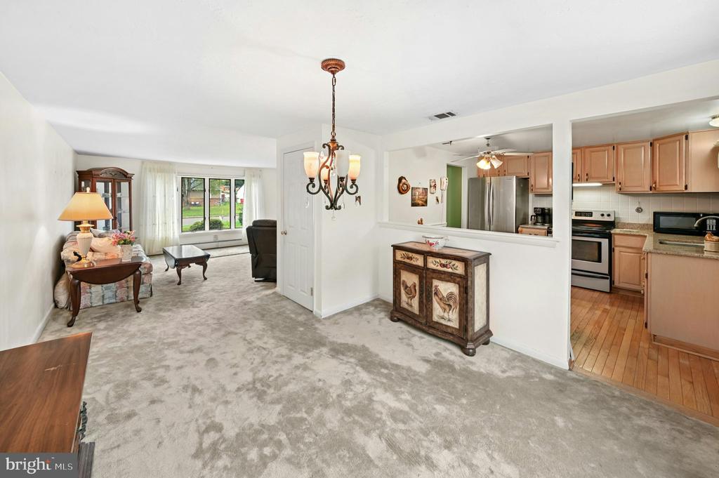 Furnished living room opening to a kitchen, with gray carpeting, a chandelier, and a large curtained window.