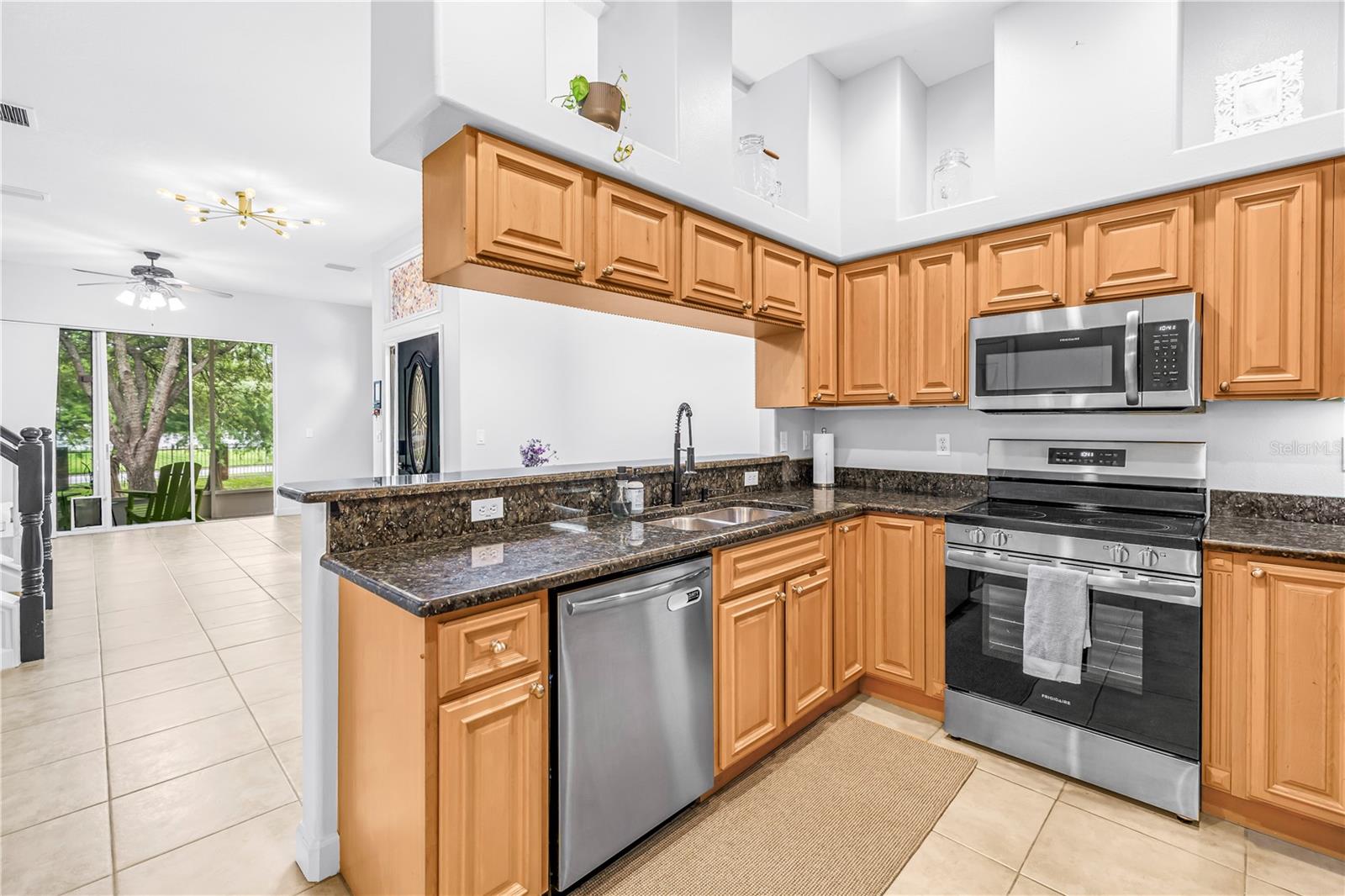 Kitchen with honey-colored cabinets, dark granite counters, stainless steel appliances, and an open view to the living area.