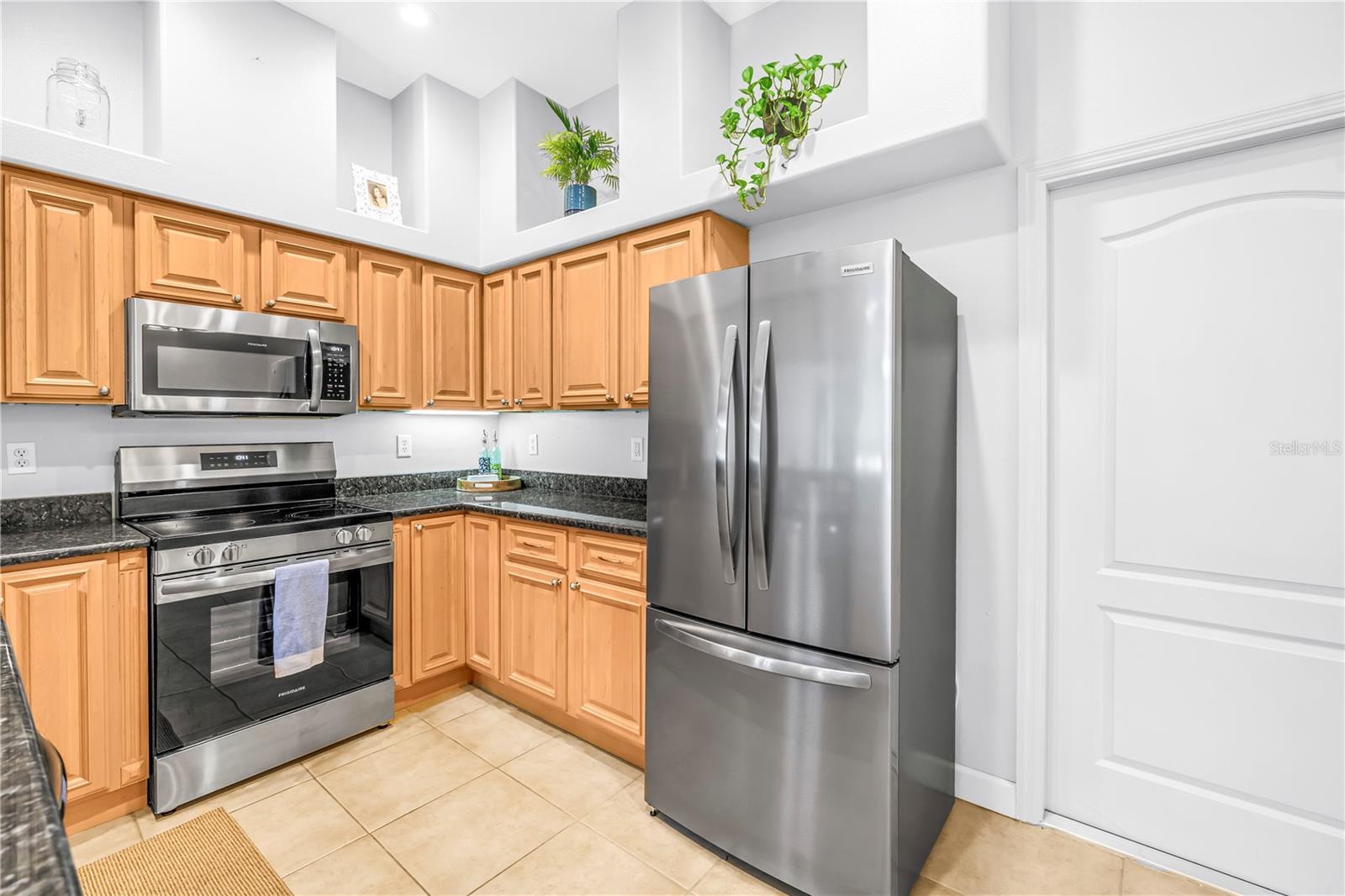 Kitchen with honey-colored cabinets, dark countertops, stainless steel appliances, and decorative plants on high shelves.