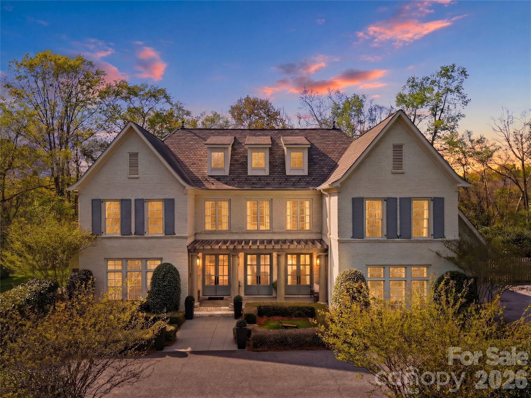 Light-brick home at dusk with three dormers, dark shutters, and a landscaped front entry.