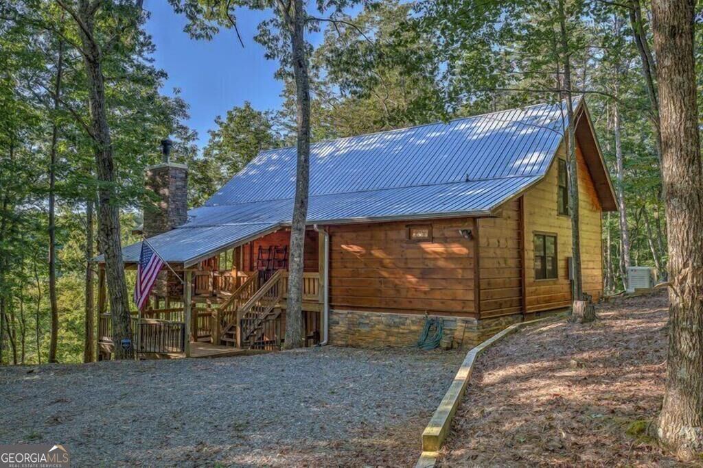 Wooded log cabin with a metal roof, covered porch, American flag, and gravel driveway.
