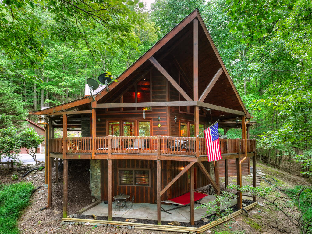 Wooden A-frame cabin in the woods with a broad deck, American flag, and red hammock.