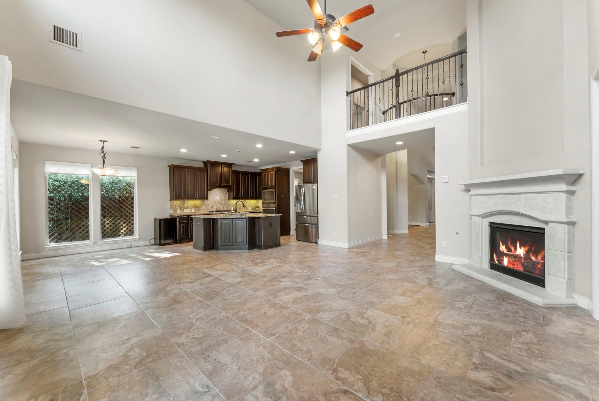 Open living room and kitchen with a tiled floor, dark cabinets, upstairs balcony, and lit fireplace.