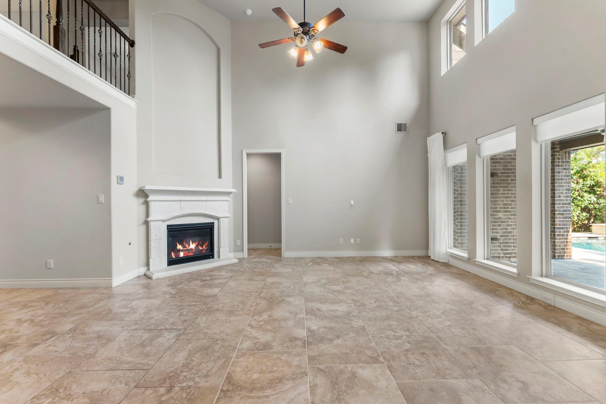 Empty living room with a vaulted ceiling, fireplace, tiled floors, and tall windows overlooking a pool.