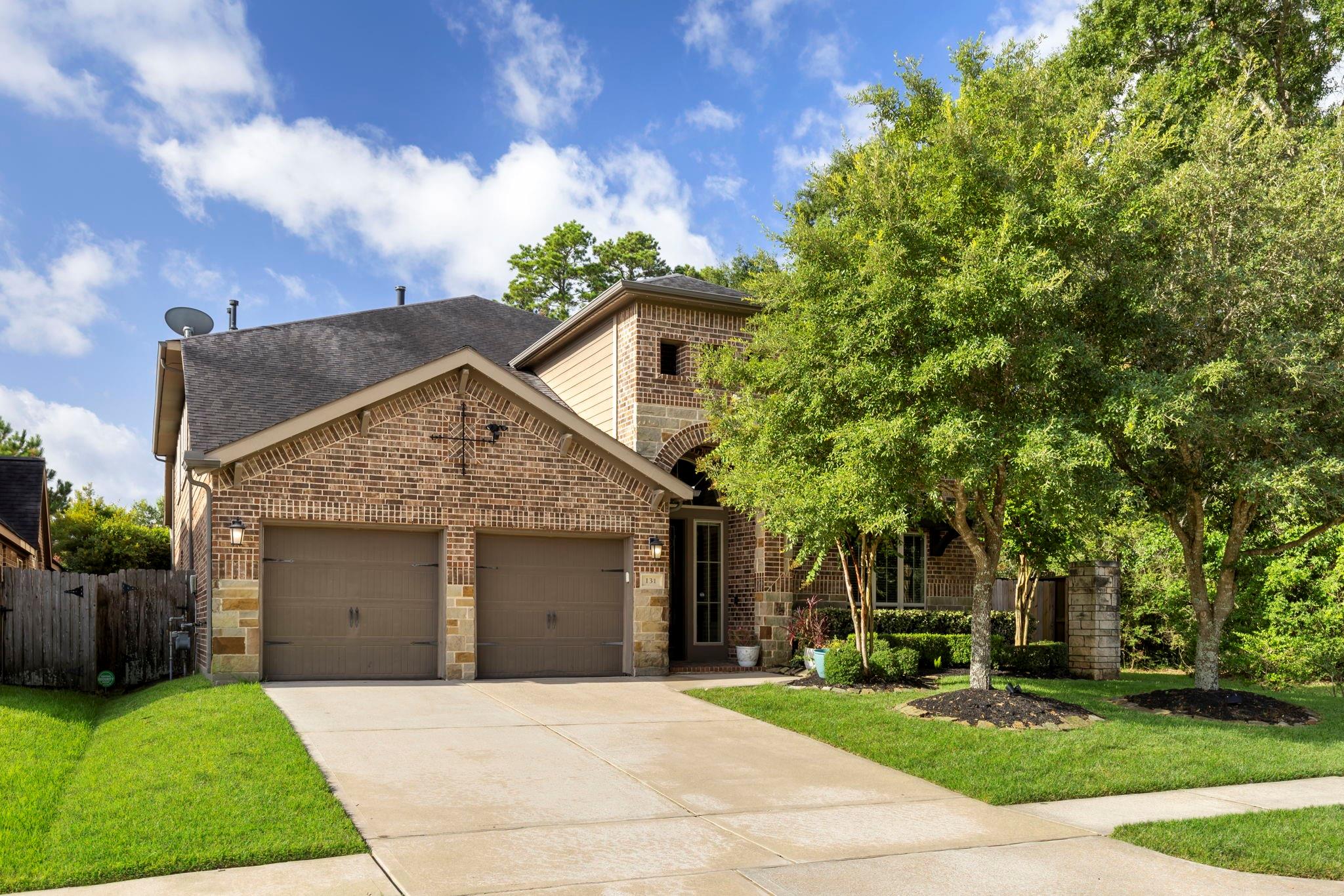 Brick home with two garage doors, a wide driveway, mature trees, and a green lawn.