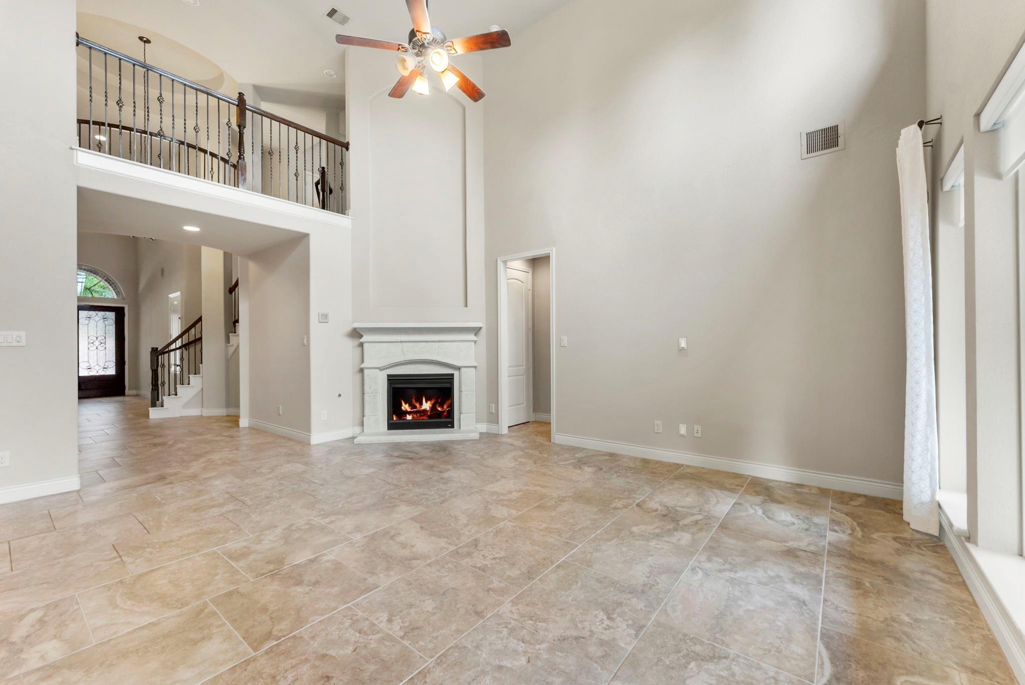 Unfurnished living room with a tall ceiling, lit fireplace, tile floors, and upstairs balcony railing.