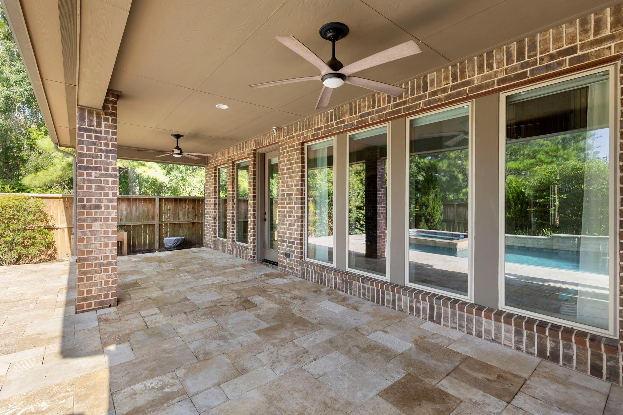 Covered brick patio with stone tile flooring, two ceiling fans, and a pool visible through the windows.