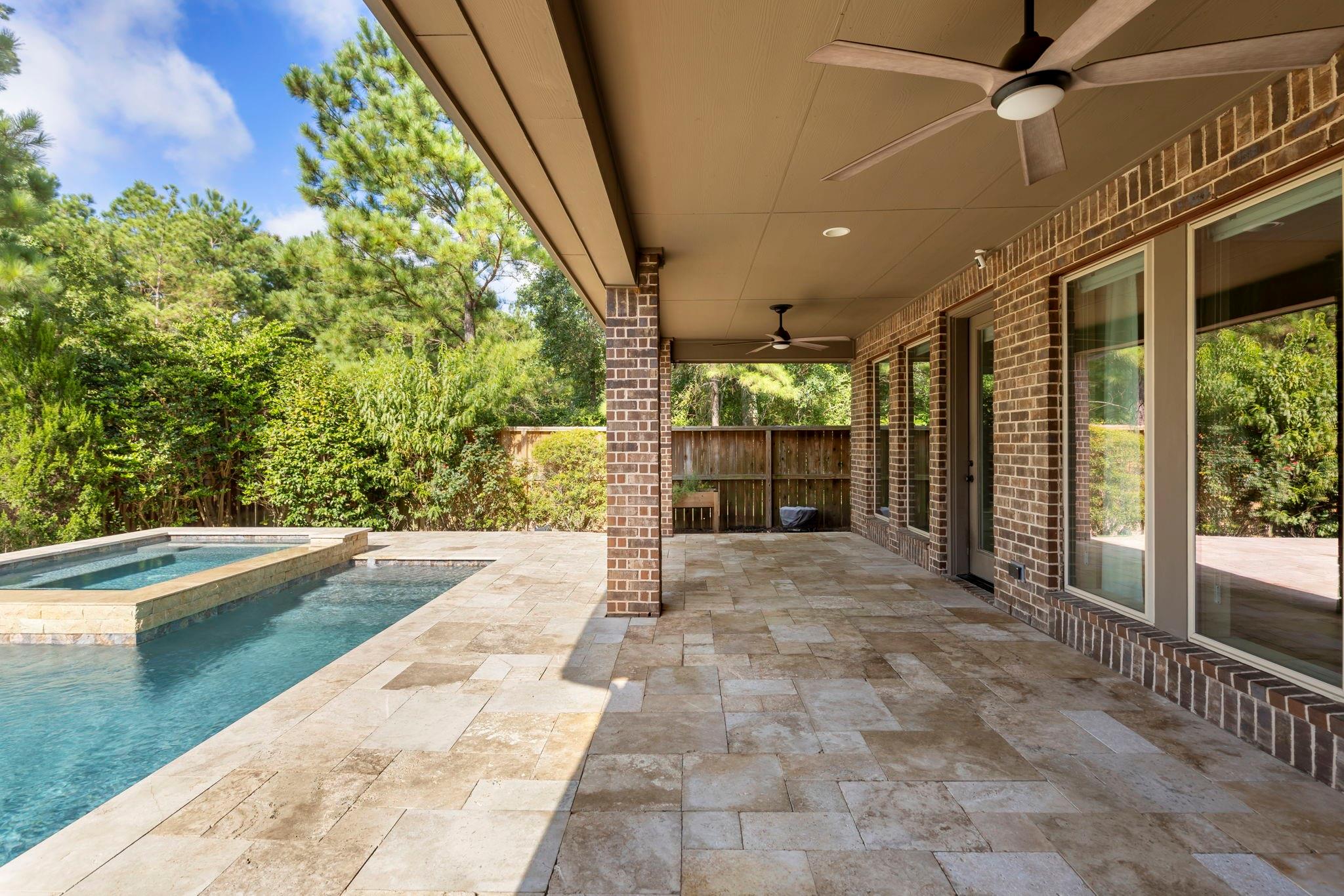 Covered brick patio beside a pool, with stone pavers and a leafy wooded backdrop.