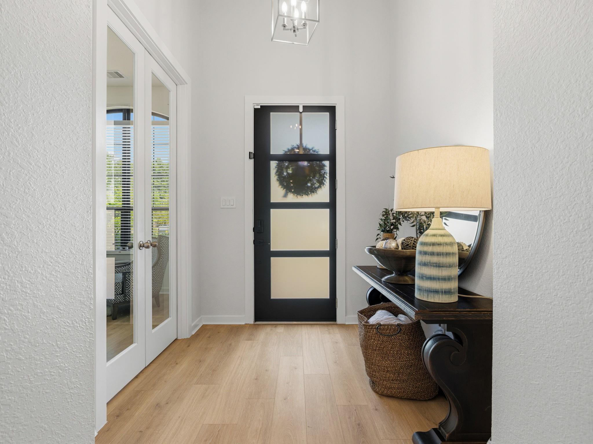 Bright entryway with a black frosted-glass front door, light wood flooring, and a console table with a large lamp.