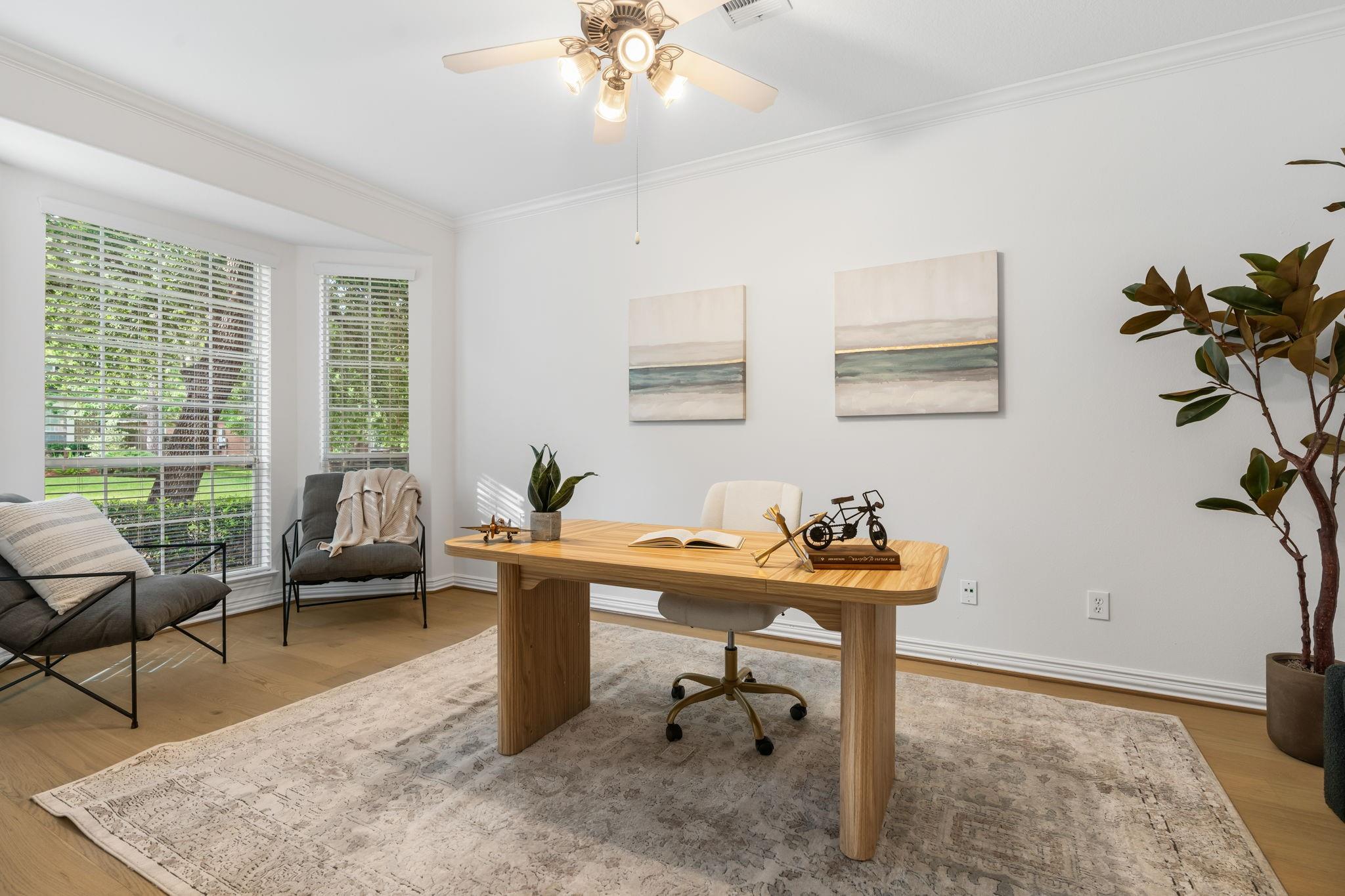 Bright home office with a wood desk, patterned rug, and tall windows overlooking greenery.