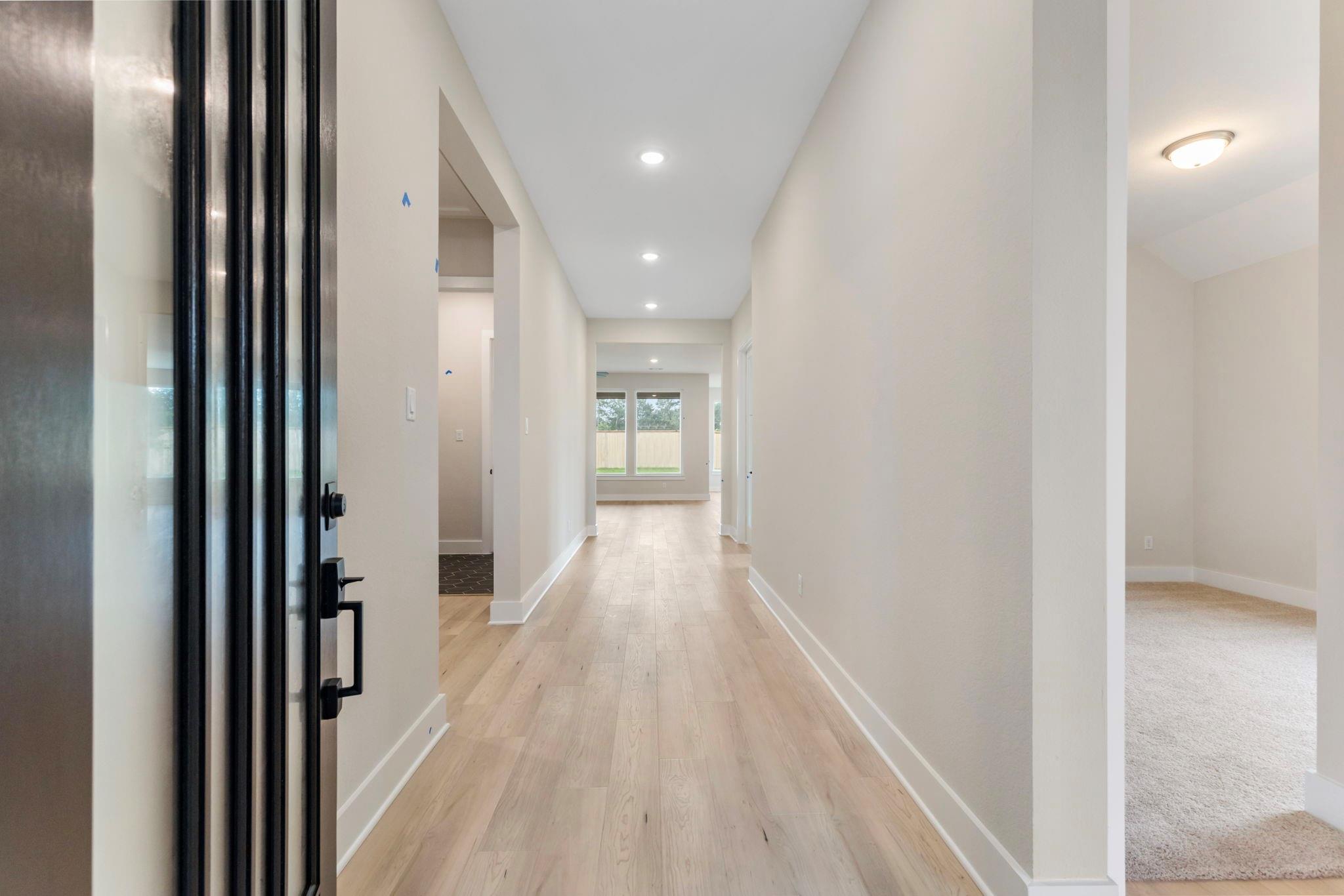 Long light-wood hallway from the black-framed glass front door, with a carpeted room on the right.