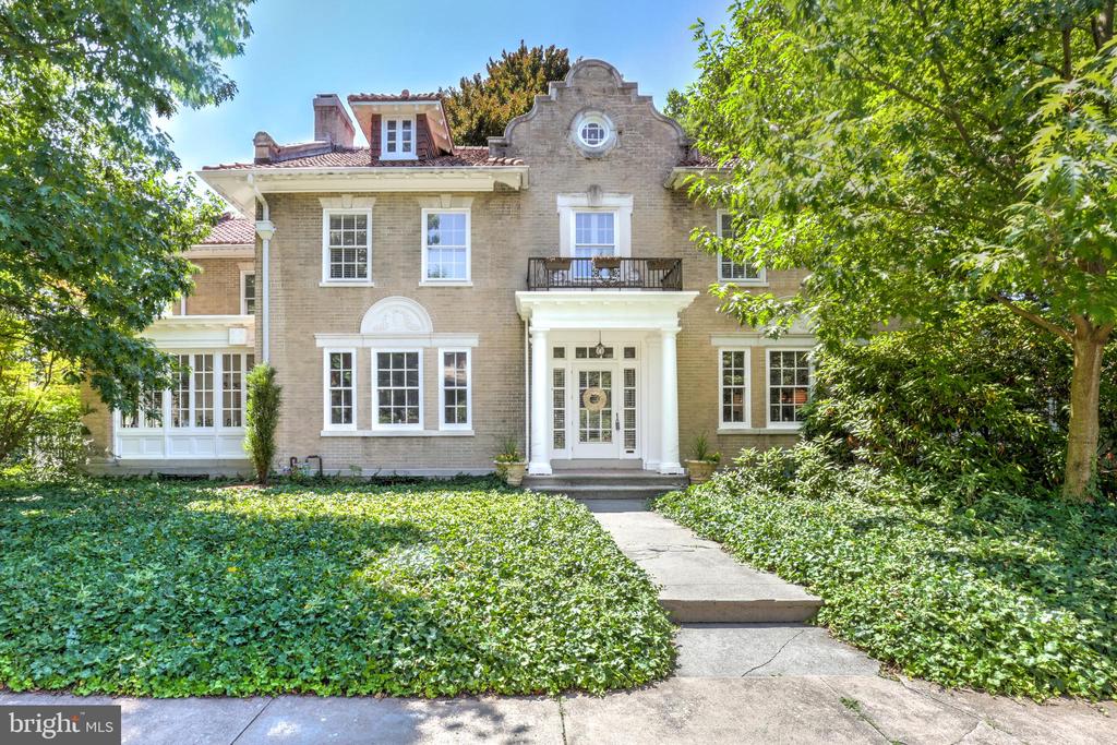 Brick house with white trim, a balcony above the entrance, and a path through leafy landscaping.