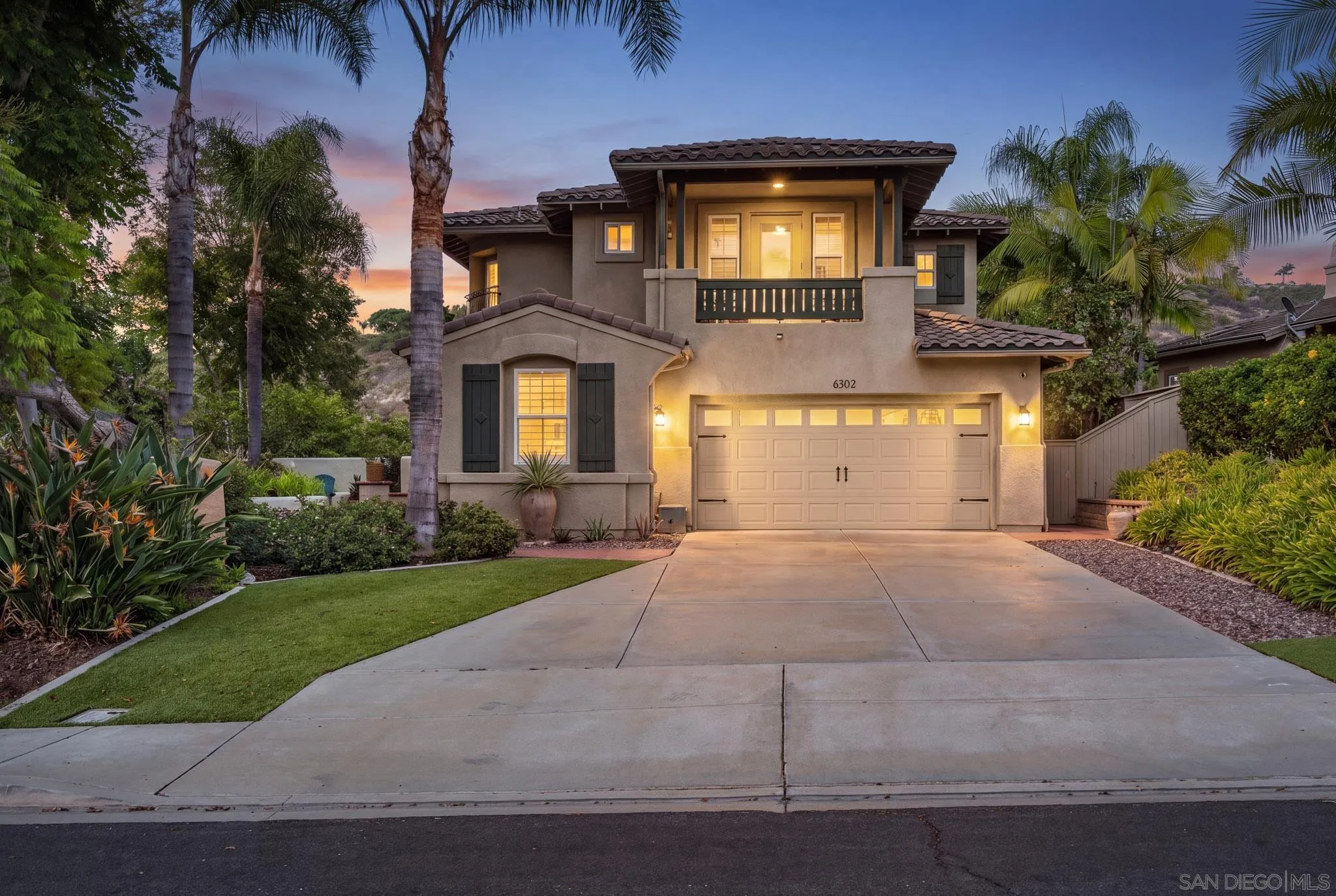 Two-story stucco home with a balcony and two-car garage, framed by palm trees at dusk.