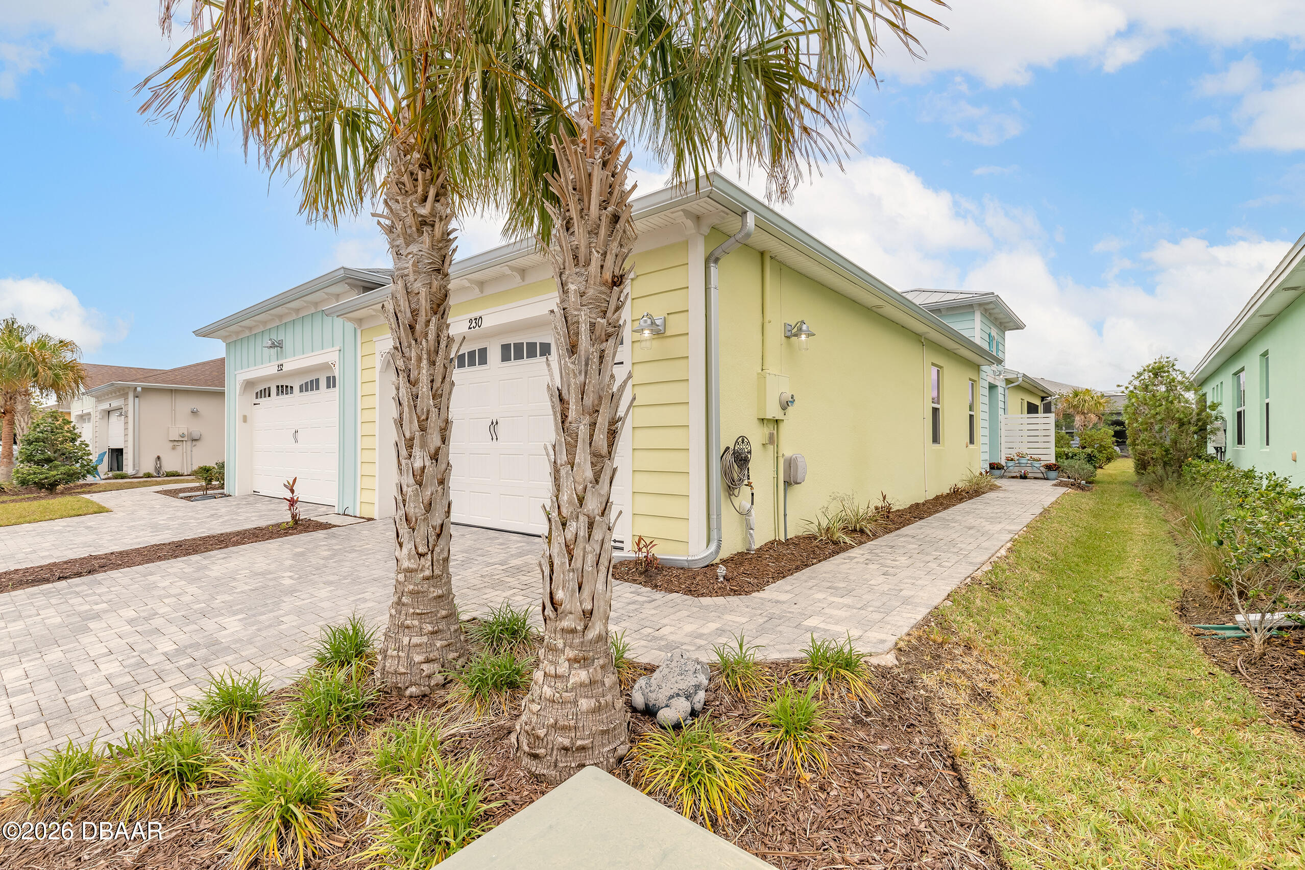 Pastel yellow coastal home with two garage doors, palm trees, and a paved walkway.