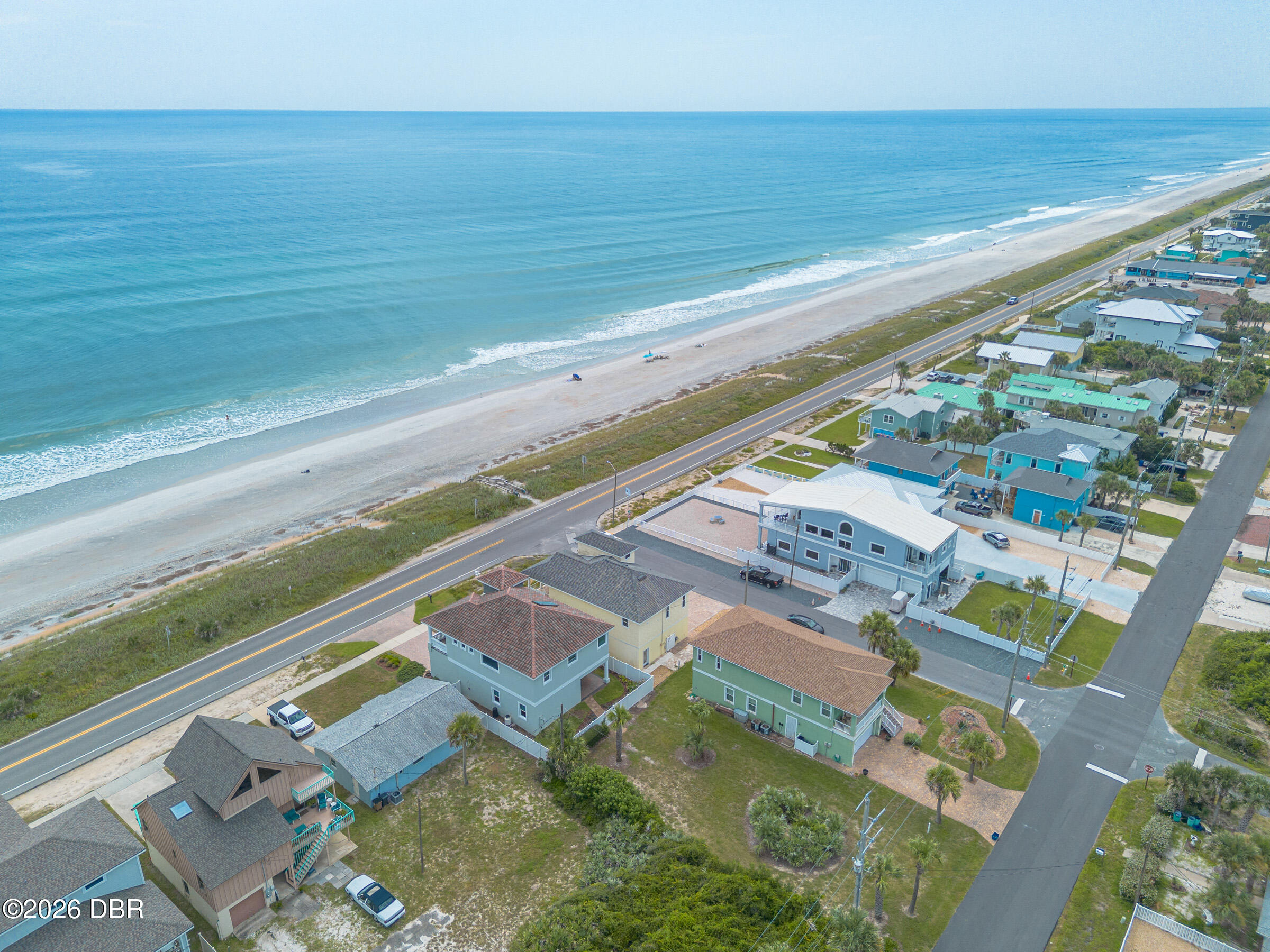 Aerial view of oceanfront homes beside a sandy beach, with a blue-gray home prominent near the coastal road.