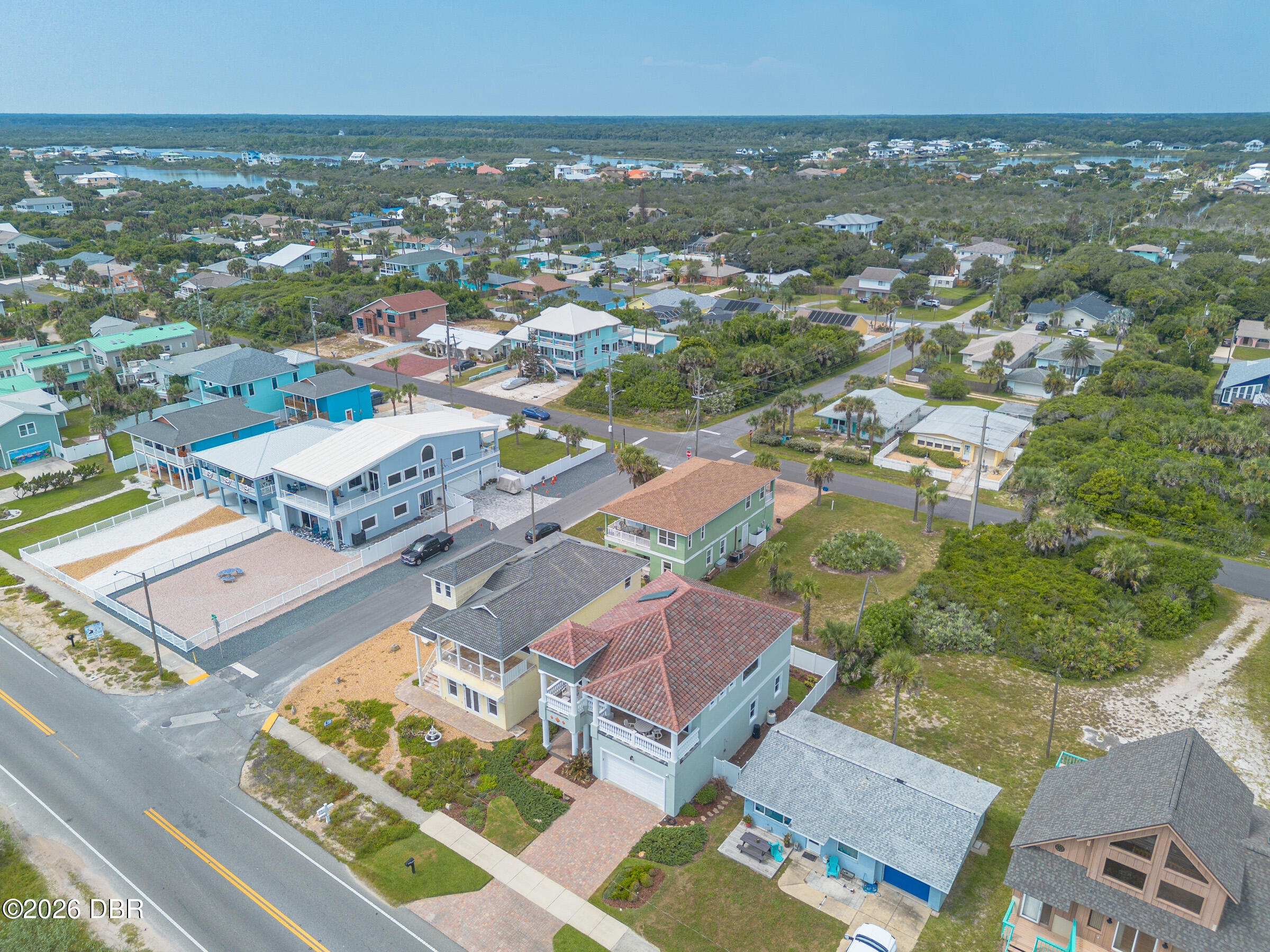 Aerial view of coastal homes, with a blue-gray multistory house and red tile roof beside the road.