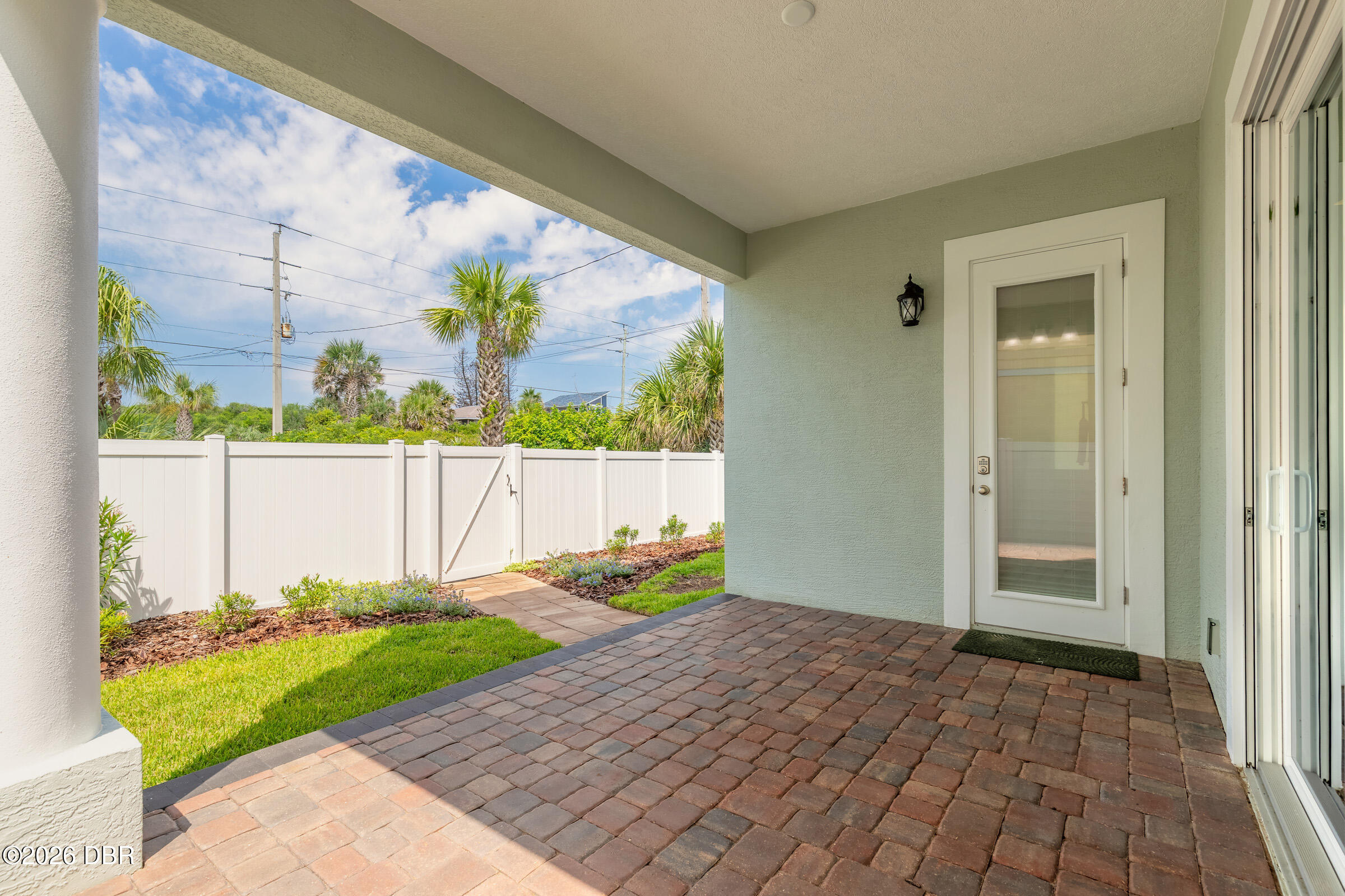 Covered brick patio beside a glass-paneled door, overlooking a fenced lawn and palm trees.