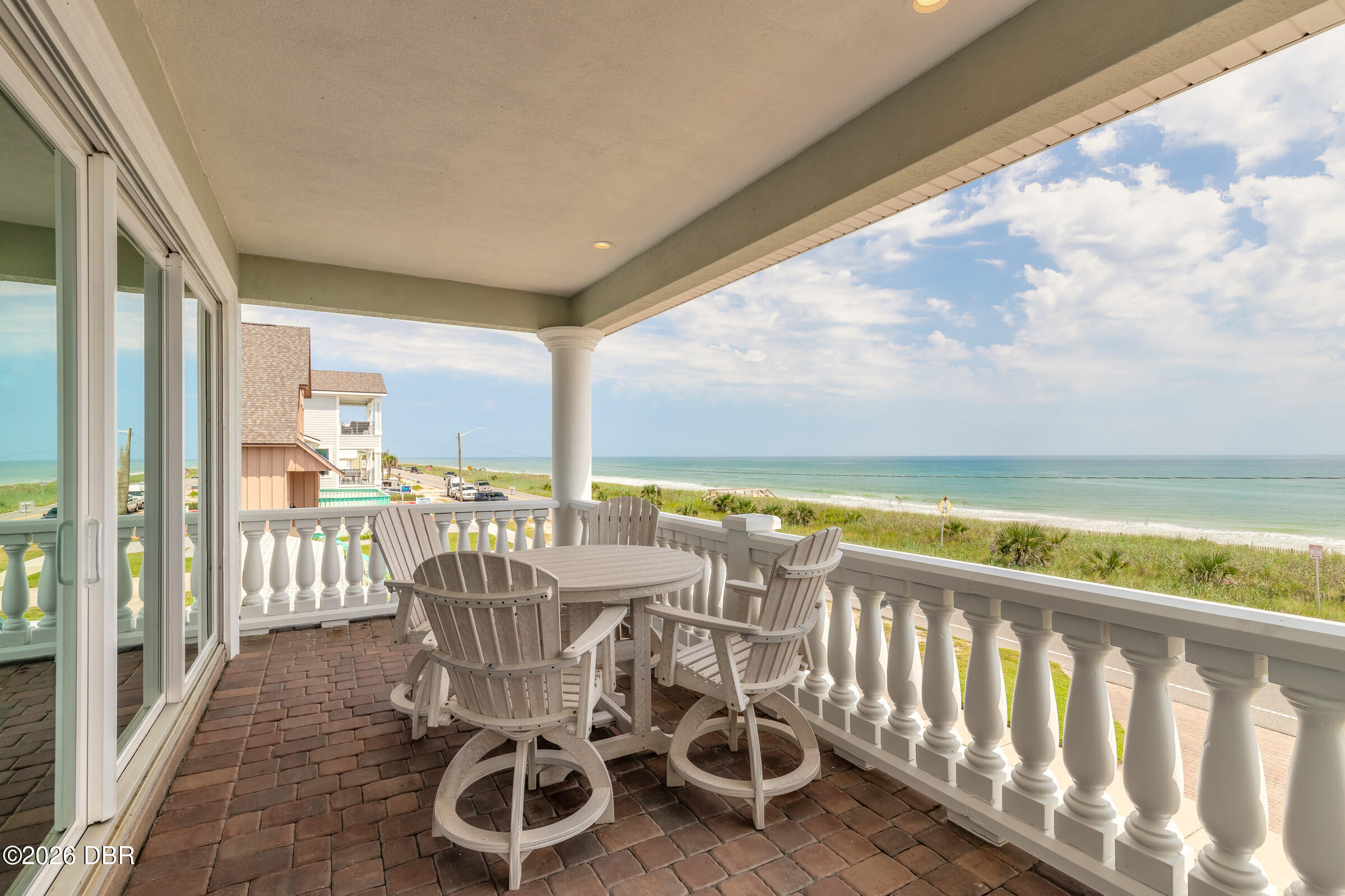 Covered oceanfront balcony with brick pavers, white railing, and outdoor dining set overlooking the beach.