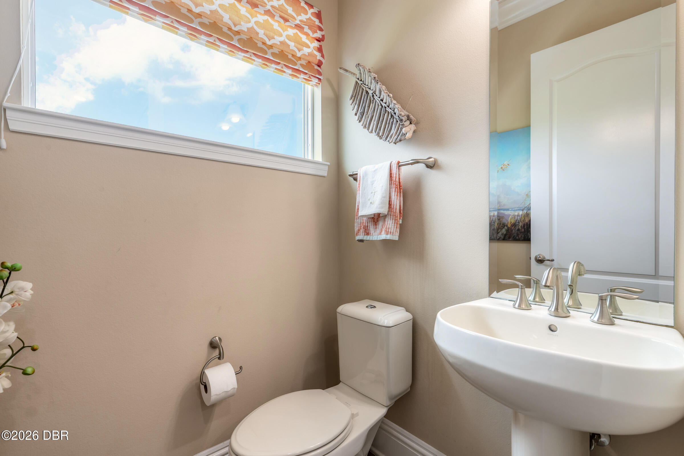 Bathroom with a pedestal sink, toilet, large window, patterned shade, and driftwood wall decor.