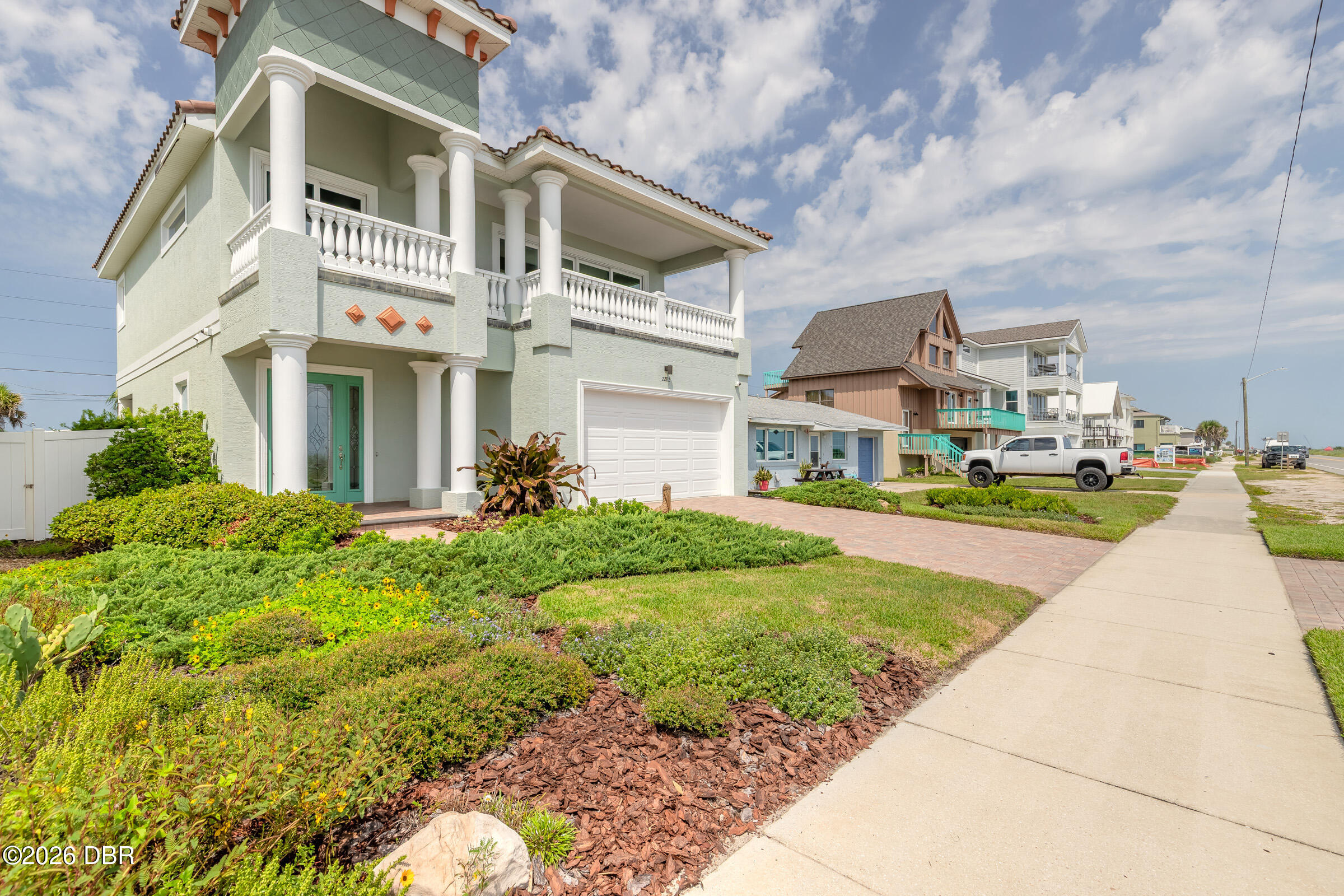 Pale green coastal home with white columns, balconies, landscaped garden, and brick driveway.