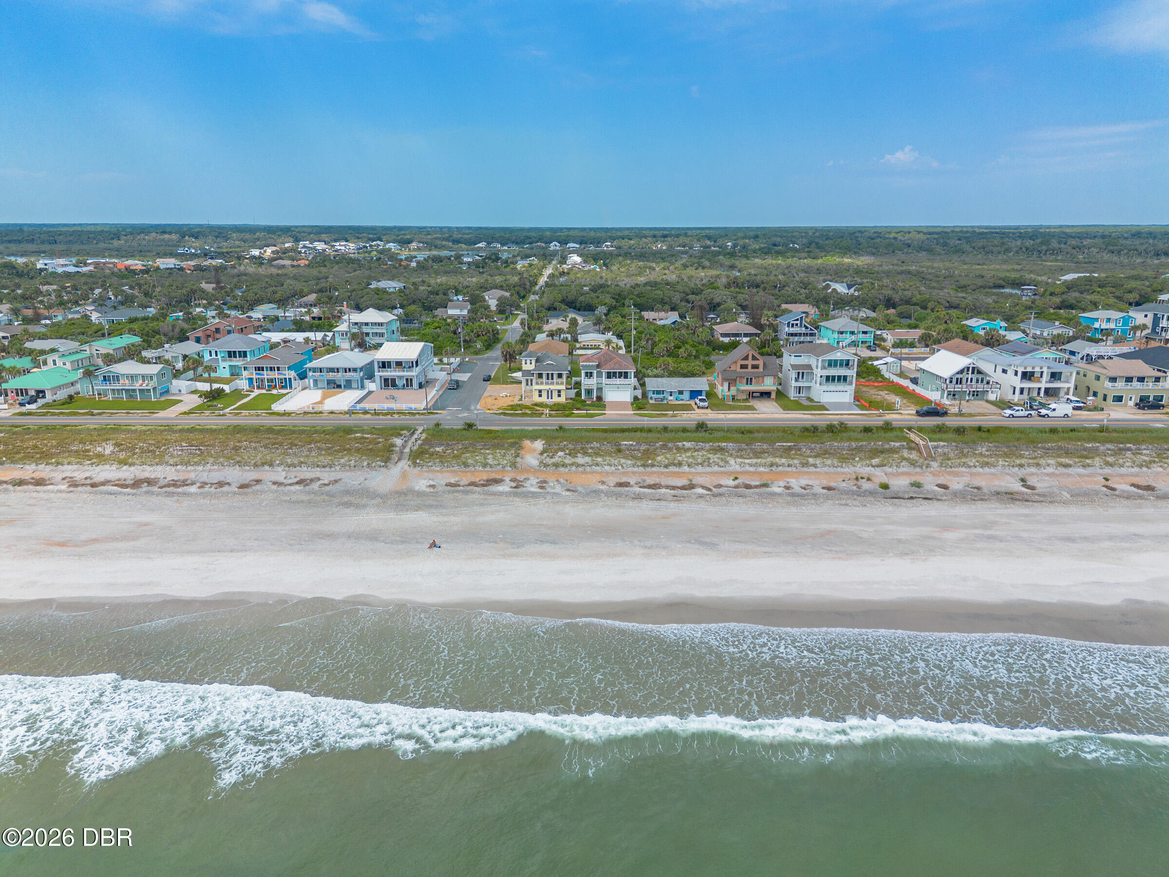 Aerial view of beachfront homes behind a wide sandy beach, with ocean waves in the foreground.