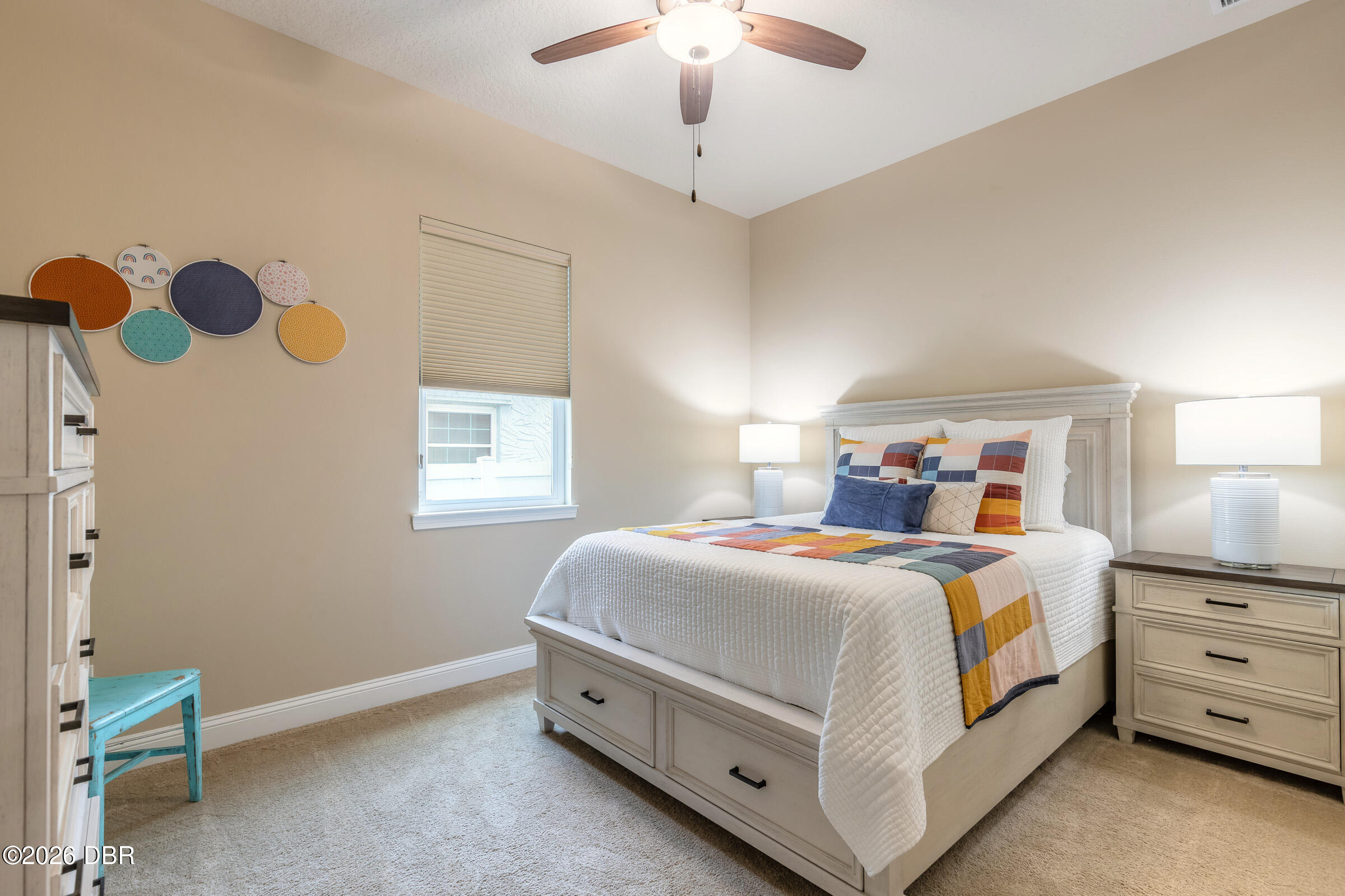 Furnished bedroom with a white bed, colorful patchwork bedding, circular wall art, and a ceiling fan.