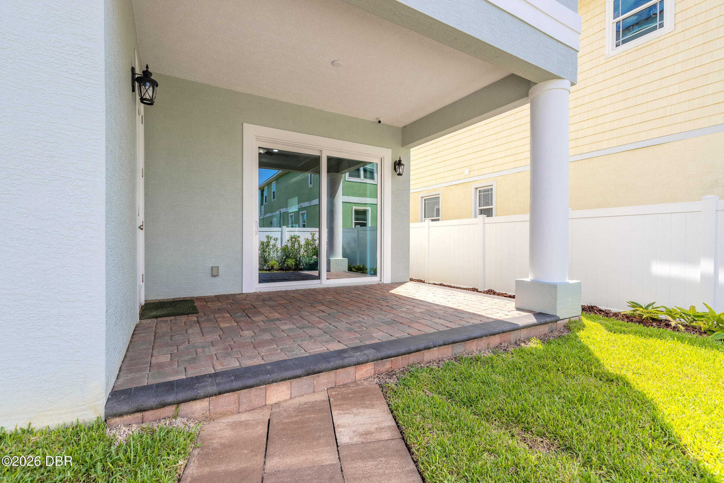 Covered rear patio with brick pavers, sliding glass doors, and a white privacy fence beside the lawn.