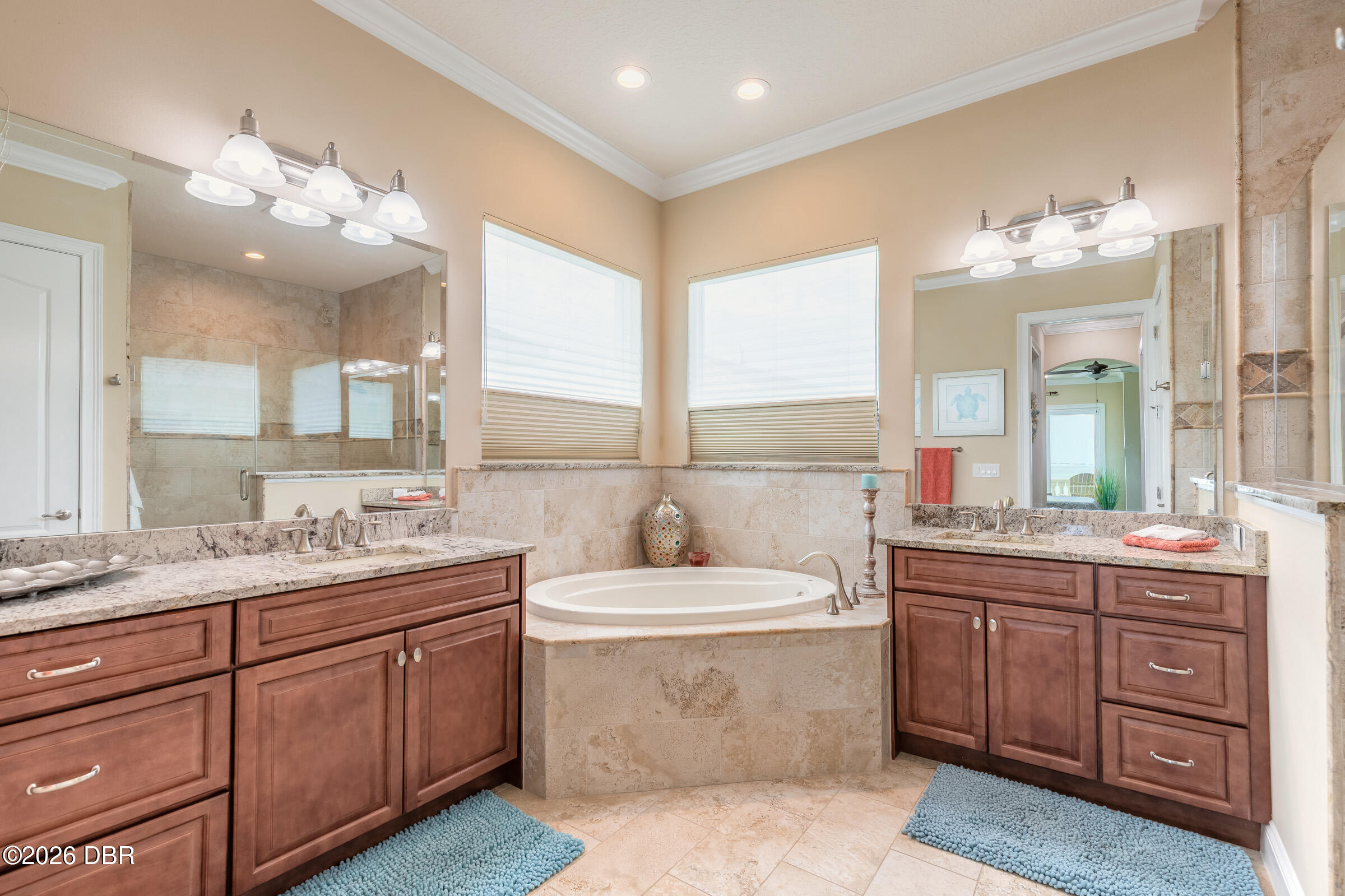 Primary bathroom with a built-in soaking tub between two wood vanities and blue bath mats.
