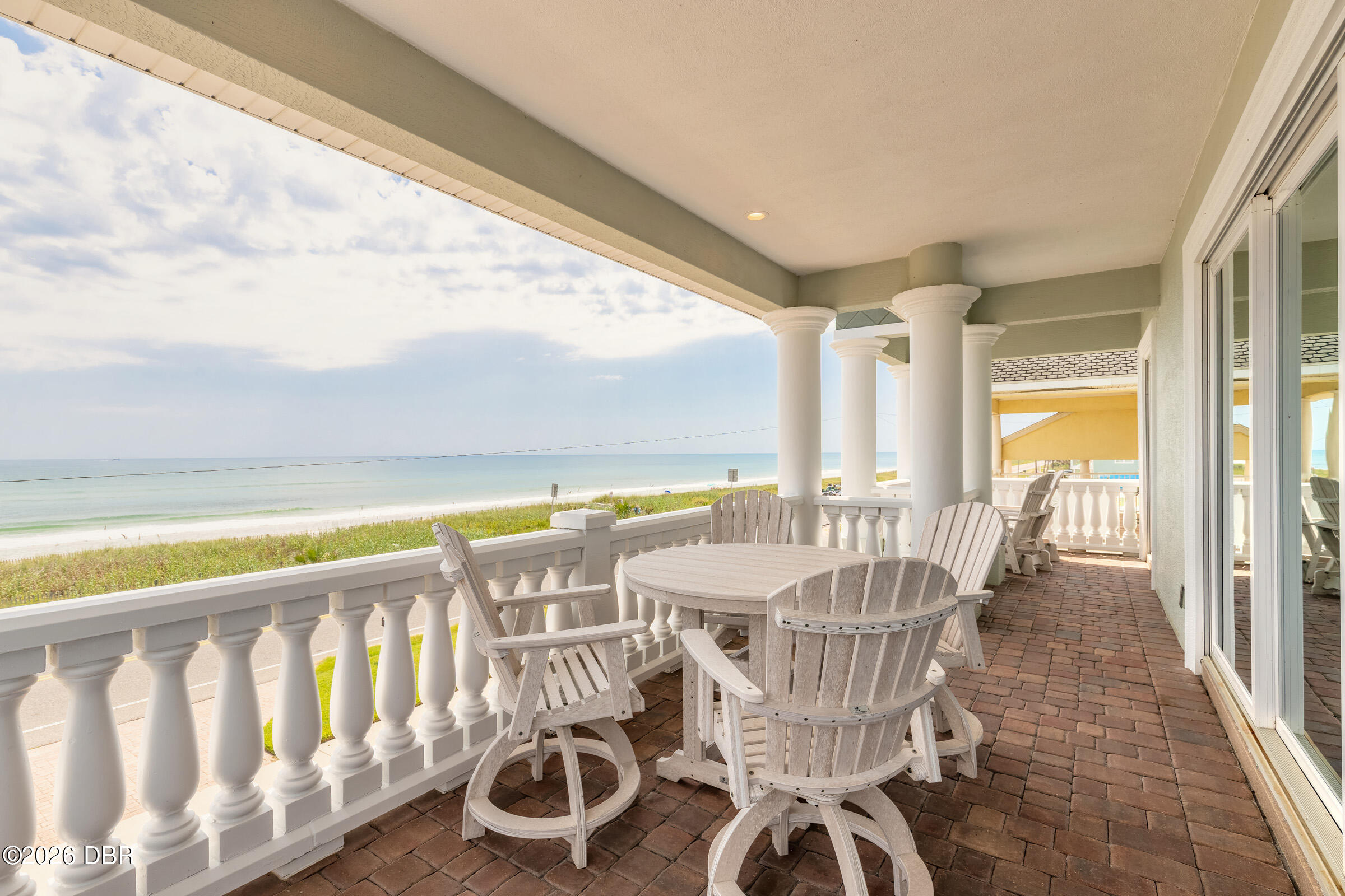 Covered oceanfront porch with white columns, brick pavers, and Adirondack seating overlooking the beach.