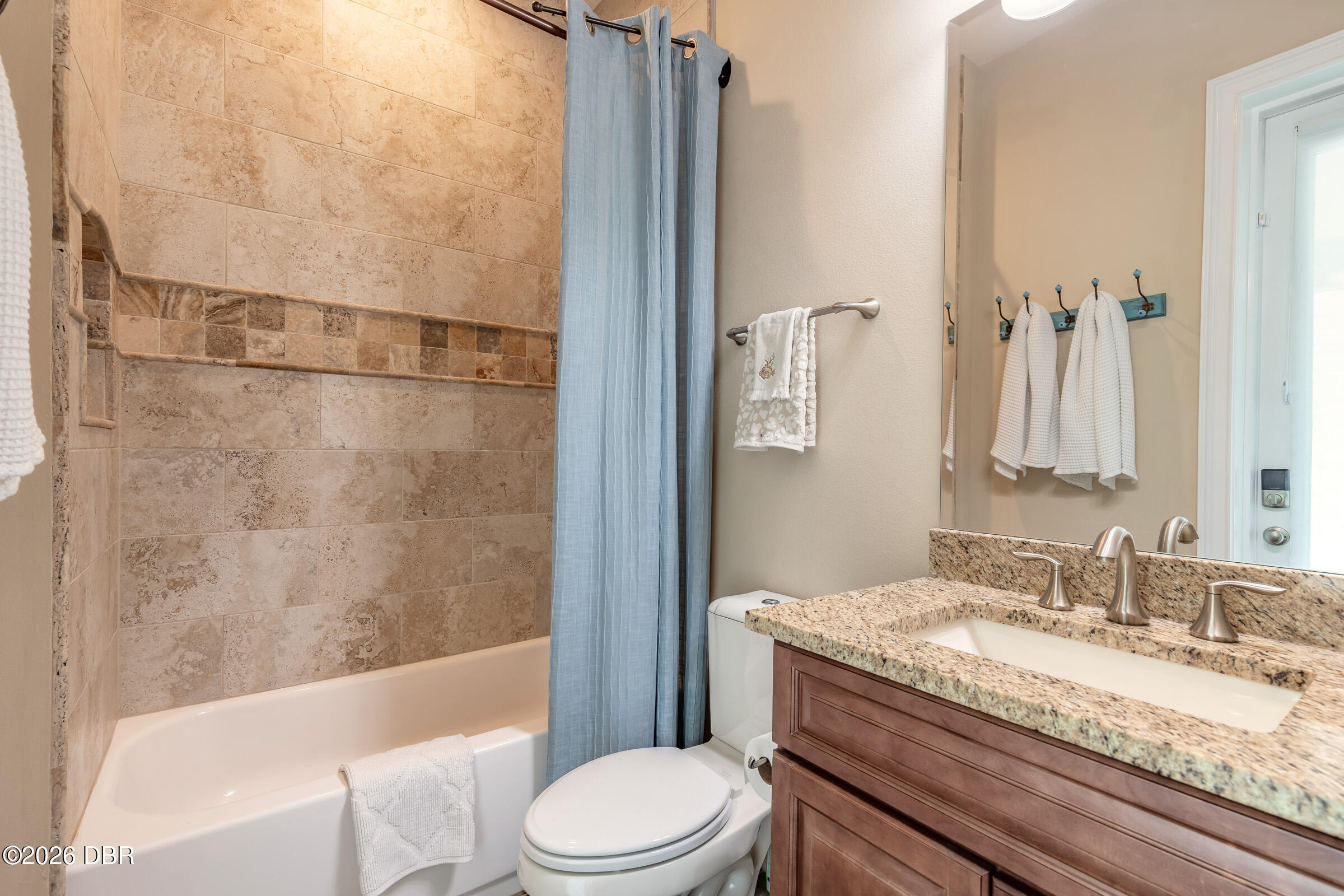 Bathroom with tan tiled tub and shower, blue curtain, and granite-topped wood vanity.