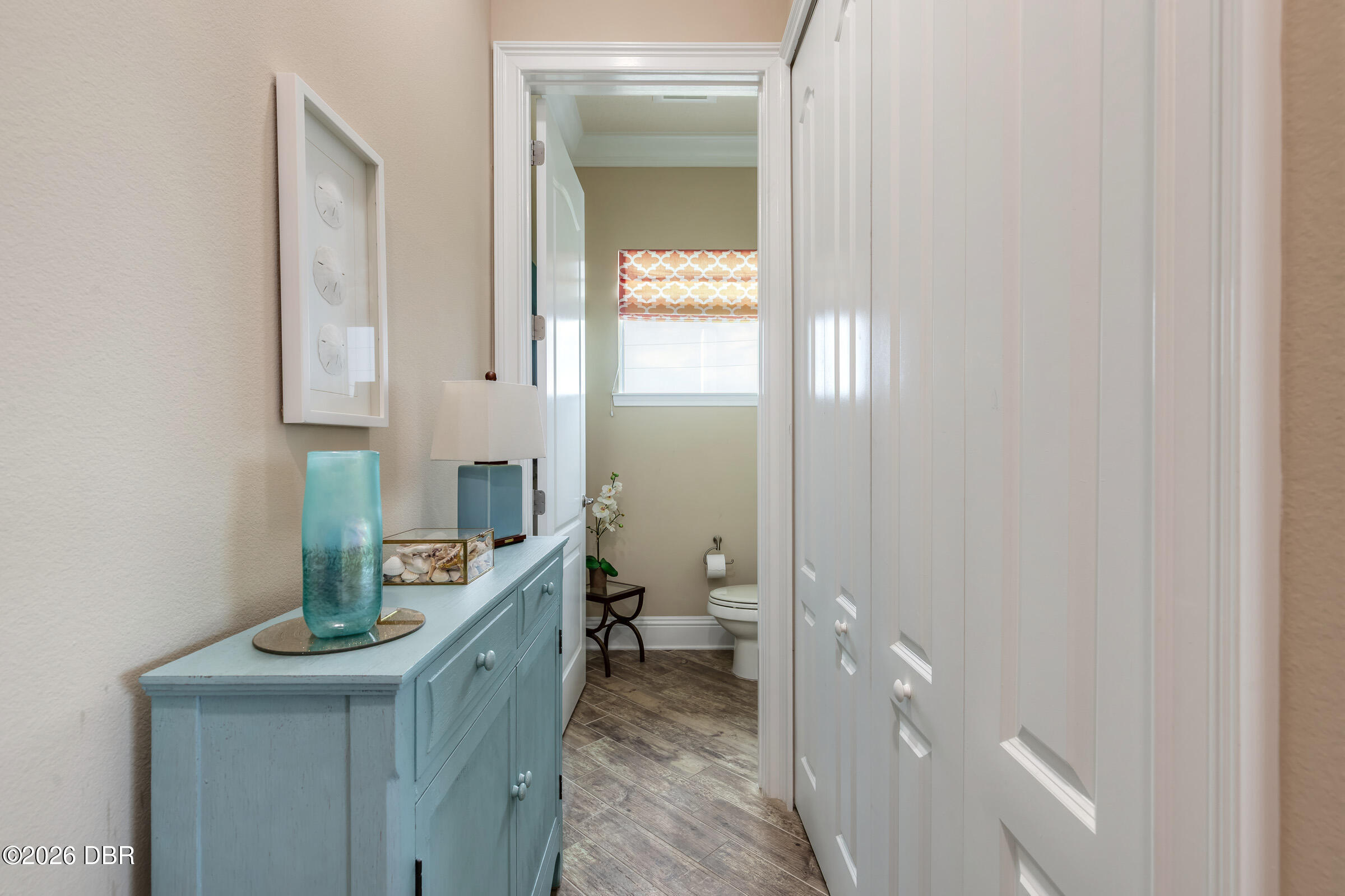 Hallway with a light blue cabinet and turquoise vase, leading to a bathroom with a patterned window shade.