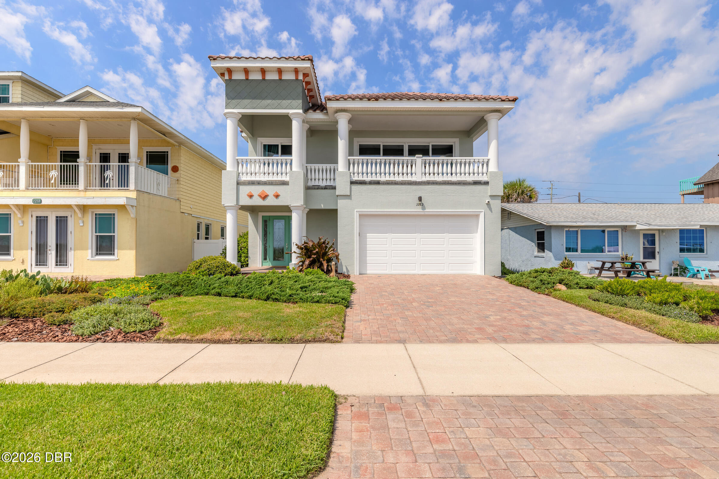 Two-story coastal home with a columned balcony, white garage, brick-paver driveway, and landscaped lawn.