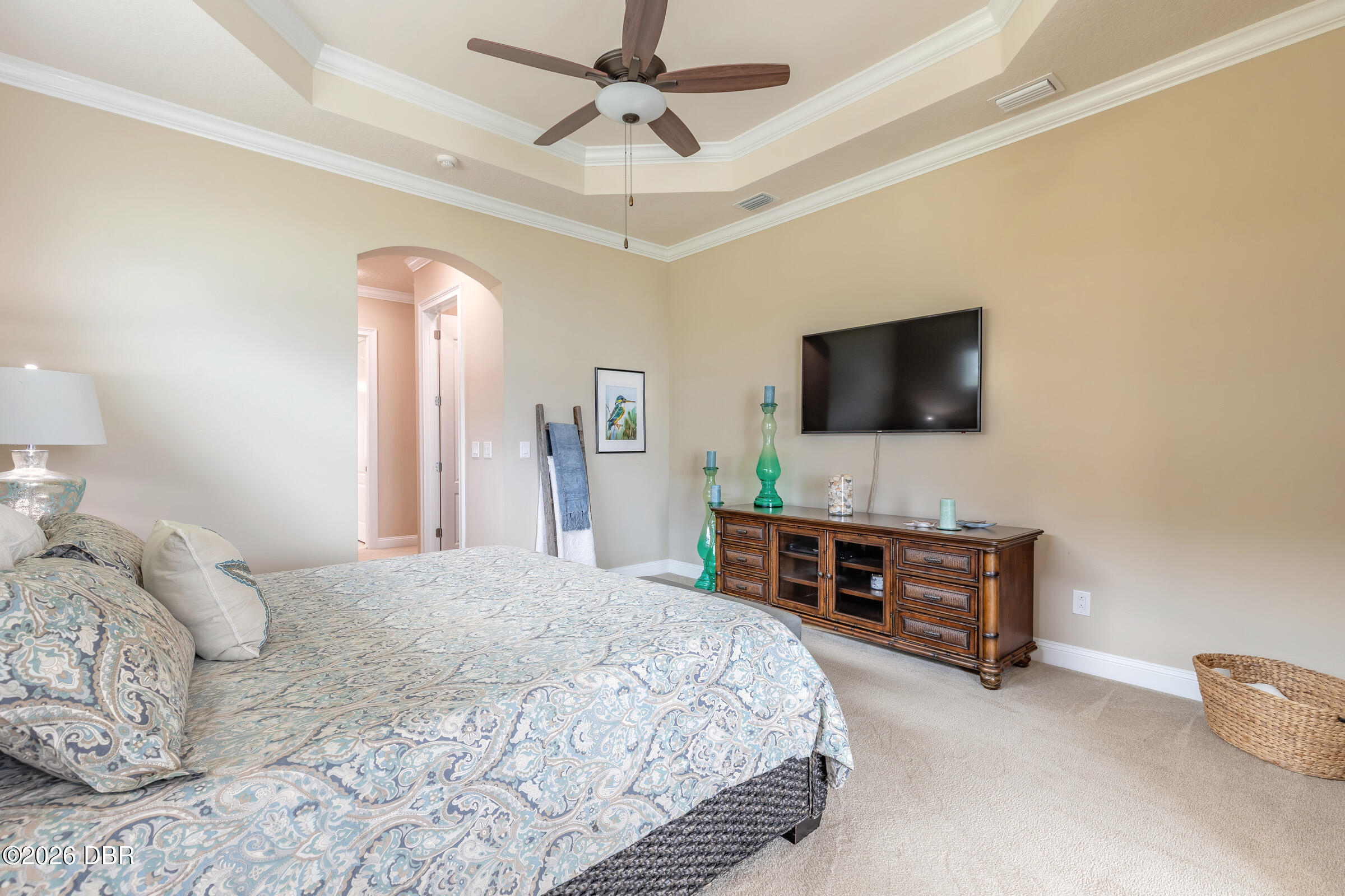 Furnished bedroom with patterned bedding, tray ceiling, ceiling fan, and television above a wooden cabinet.
