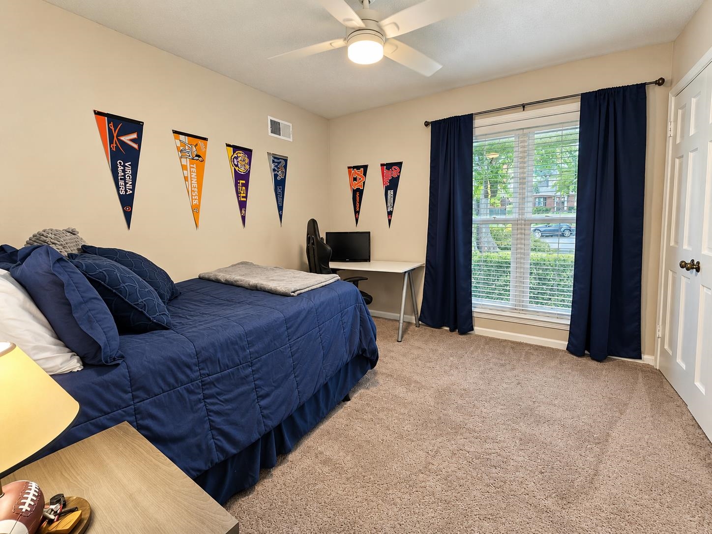 Bedroom with a navy bed, college pennants, and a desk beside a curtained window.