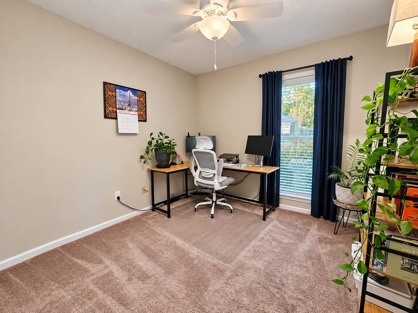 Home office with a desk and white chair beside a tall window framed by dark curtains.
