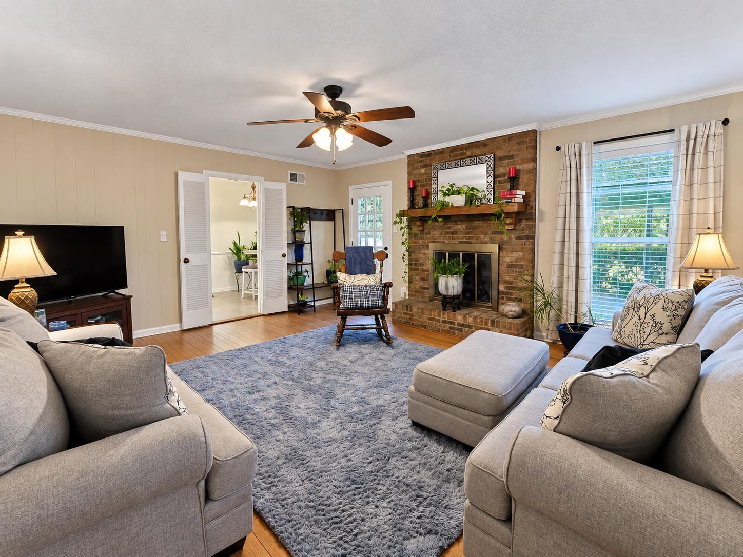 Living room with a brick fireplace, light sofas, a gray rug, and a large curtained window.