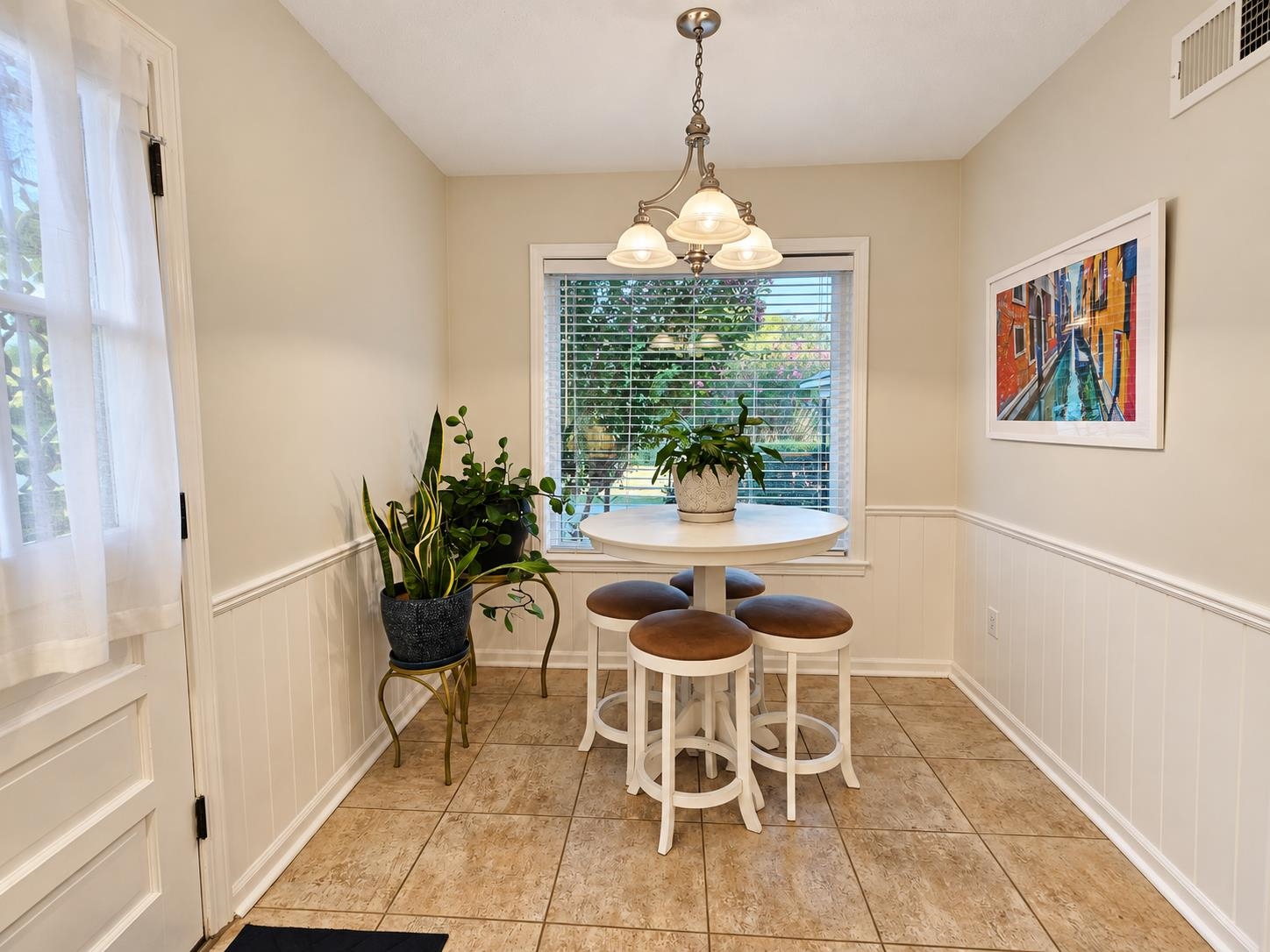 Breakfast nook with a round white table, brown-seat stools, leafy plants, and a large window with blinds.
