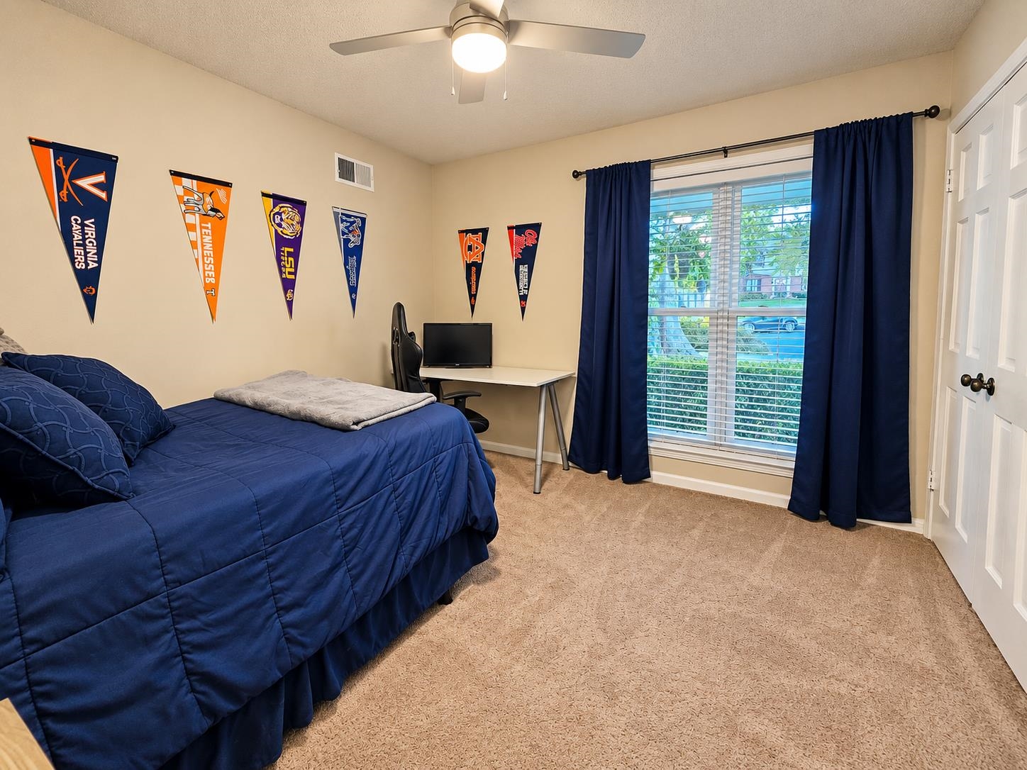 Furnished bedroom with a blue bedspread, college pennants, desk, and navy curtains.