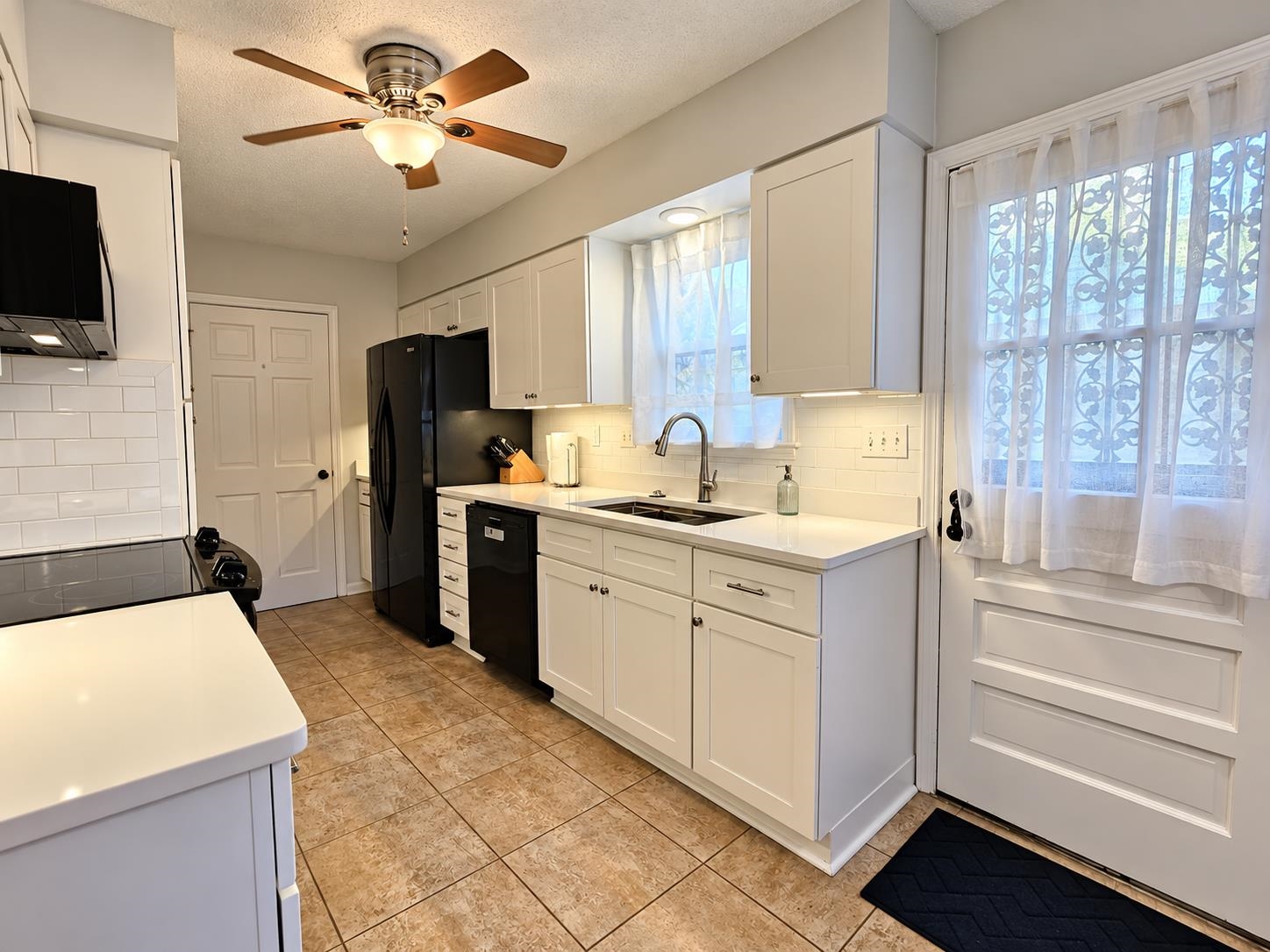 Kitchen with white cabinets, black appliances, a stainless sink, and daylight through curtained windows.